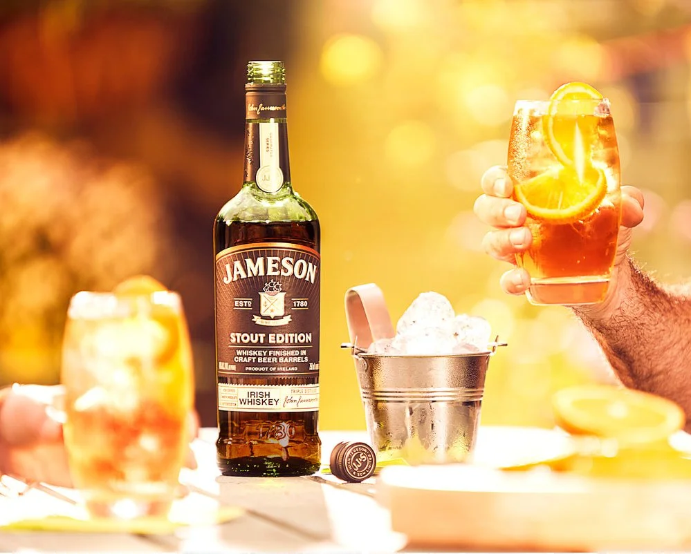 Commercial promotional photo of a bottle of Jameson Irish Whiskey Stout Edition, a glass of whiskey with ice and lemon slices, and a metal ice bucket with ice cubes on a table outdoors shooting in location with high quality beautiful lighting.