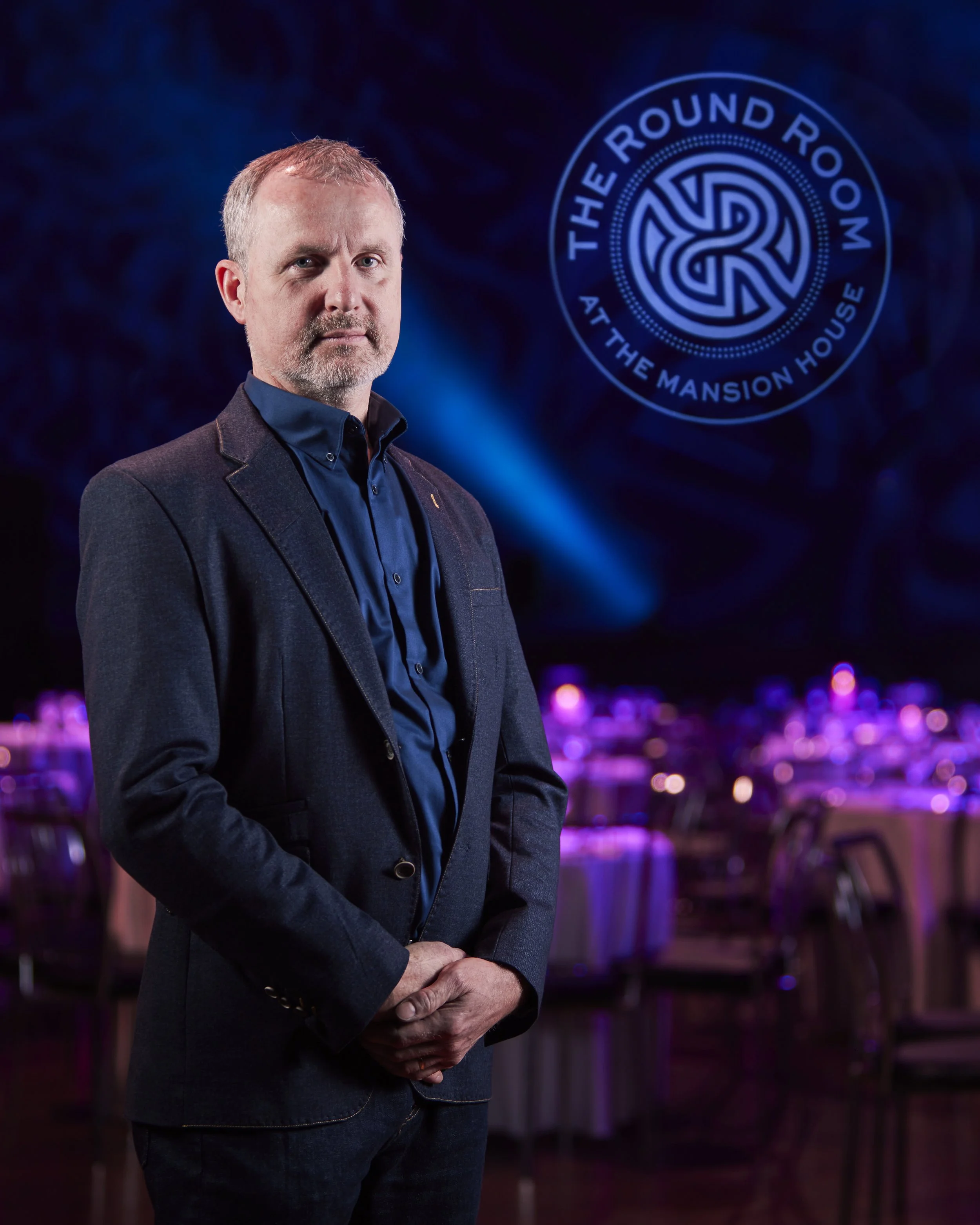 Corporate portrait photography of a man with short light brown hair and a beard, wearing a dark suit jacket and blue shirt, standing in a dimly lit event space with purple lighting and round tables in the background. 