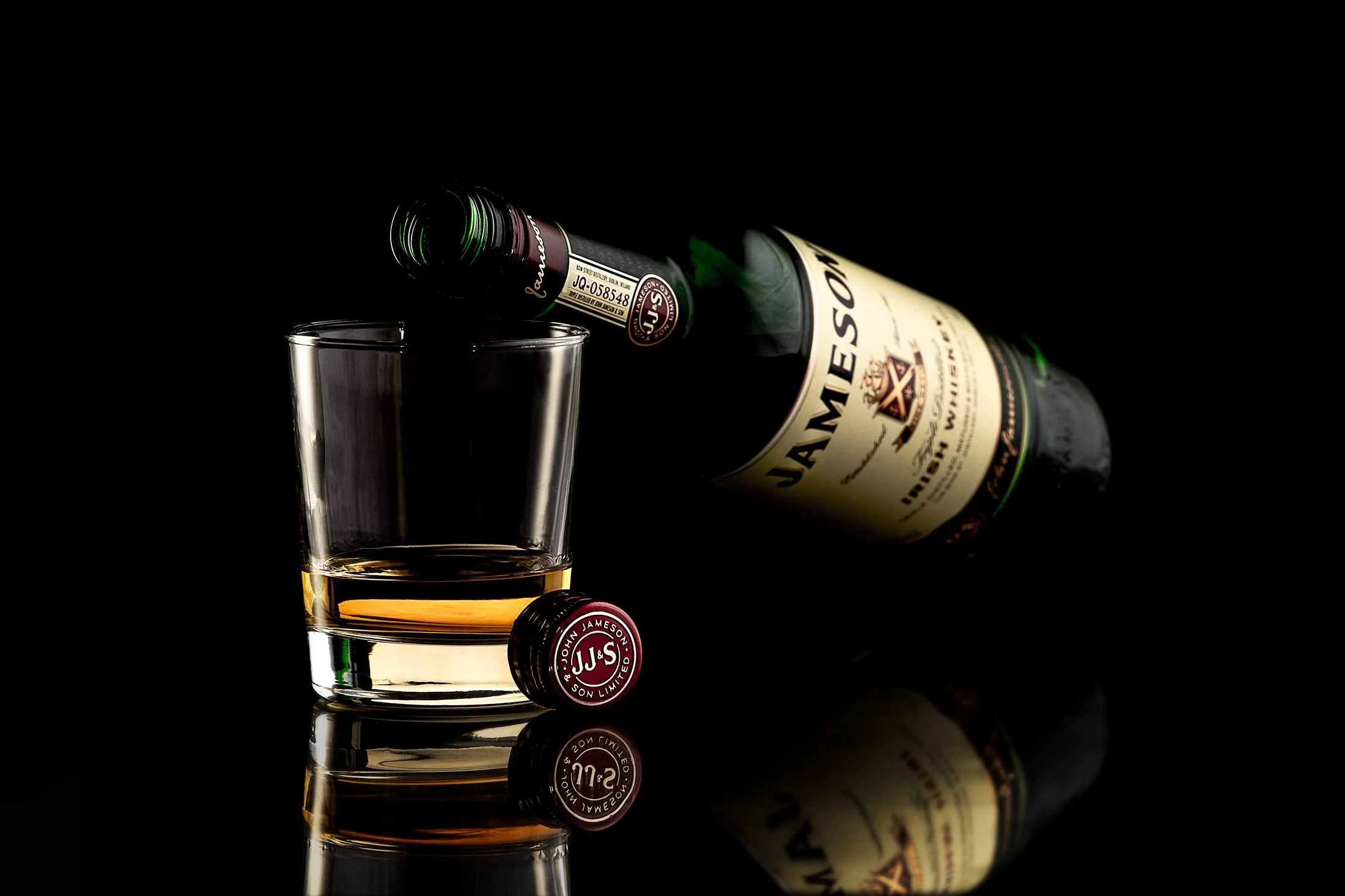 Advertising profession photo taken in studio of a bottle of Jameson Irish whiskey lying on its side, a glass of whiskey with a small amount poured in, and the bottle's cork on a reflective surface against a black background.