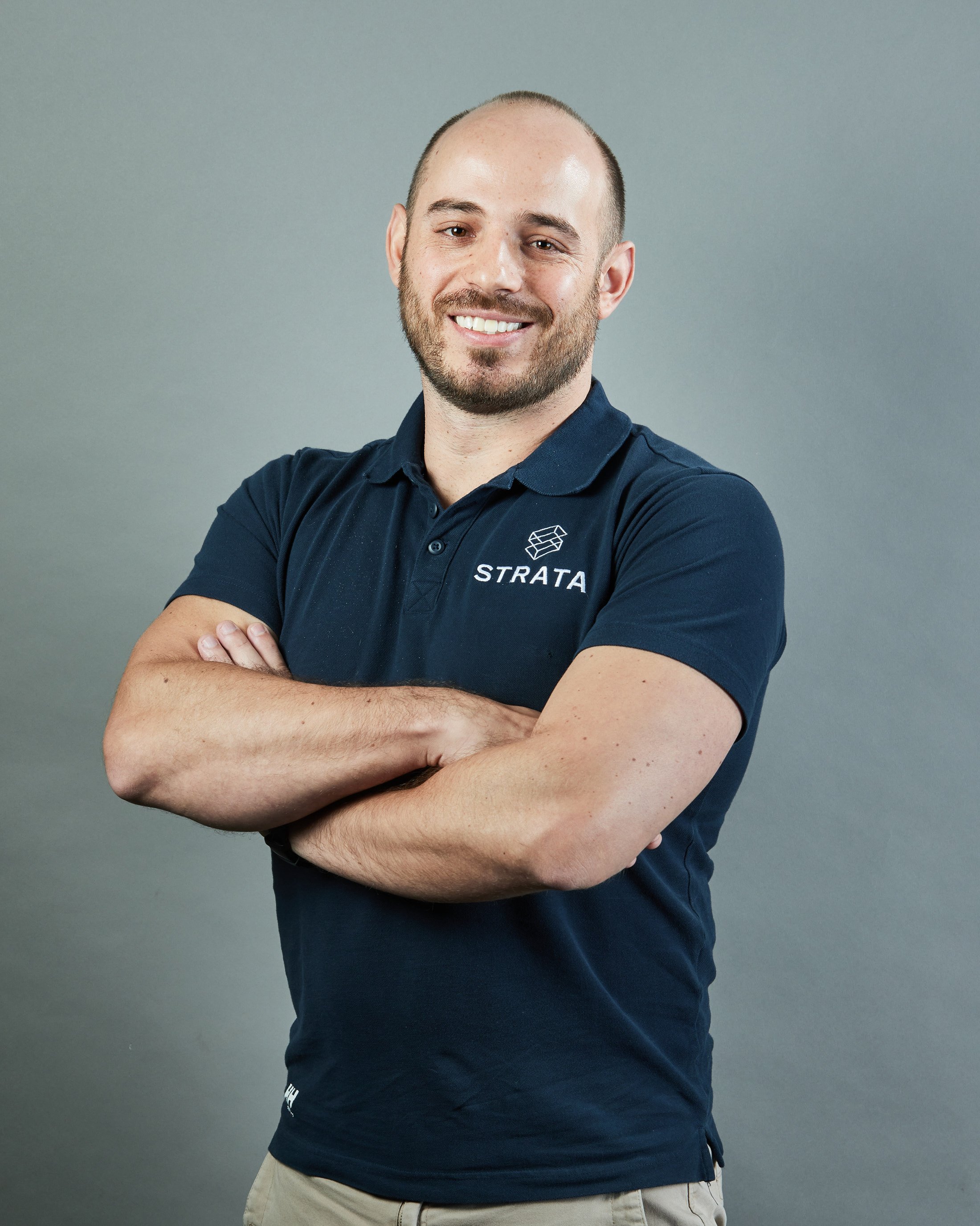 A smiling employe man with a beard and short hair, wearing a navy polo shirt with the logo and name "STRATA," standing with arms crossed against a plain gray background with profesional high quality photography lighting in location.