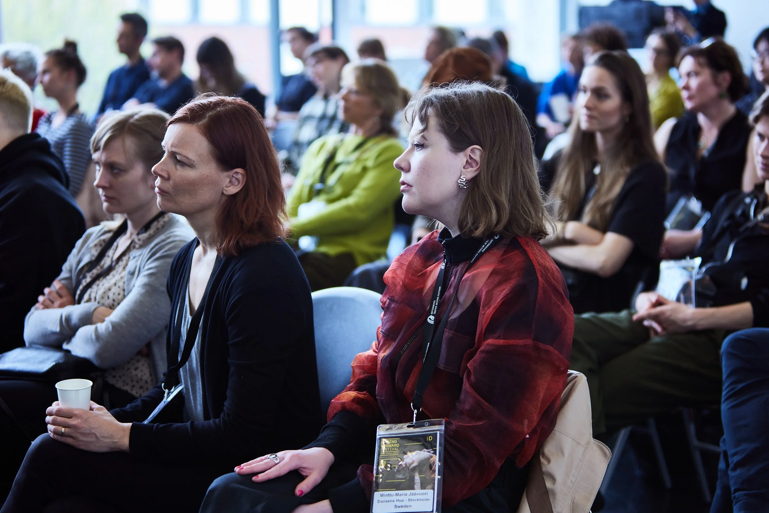 Audience attending a conference, sitting attentively with some taking notes or holding papers, in a bright room with large windows.