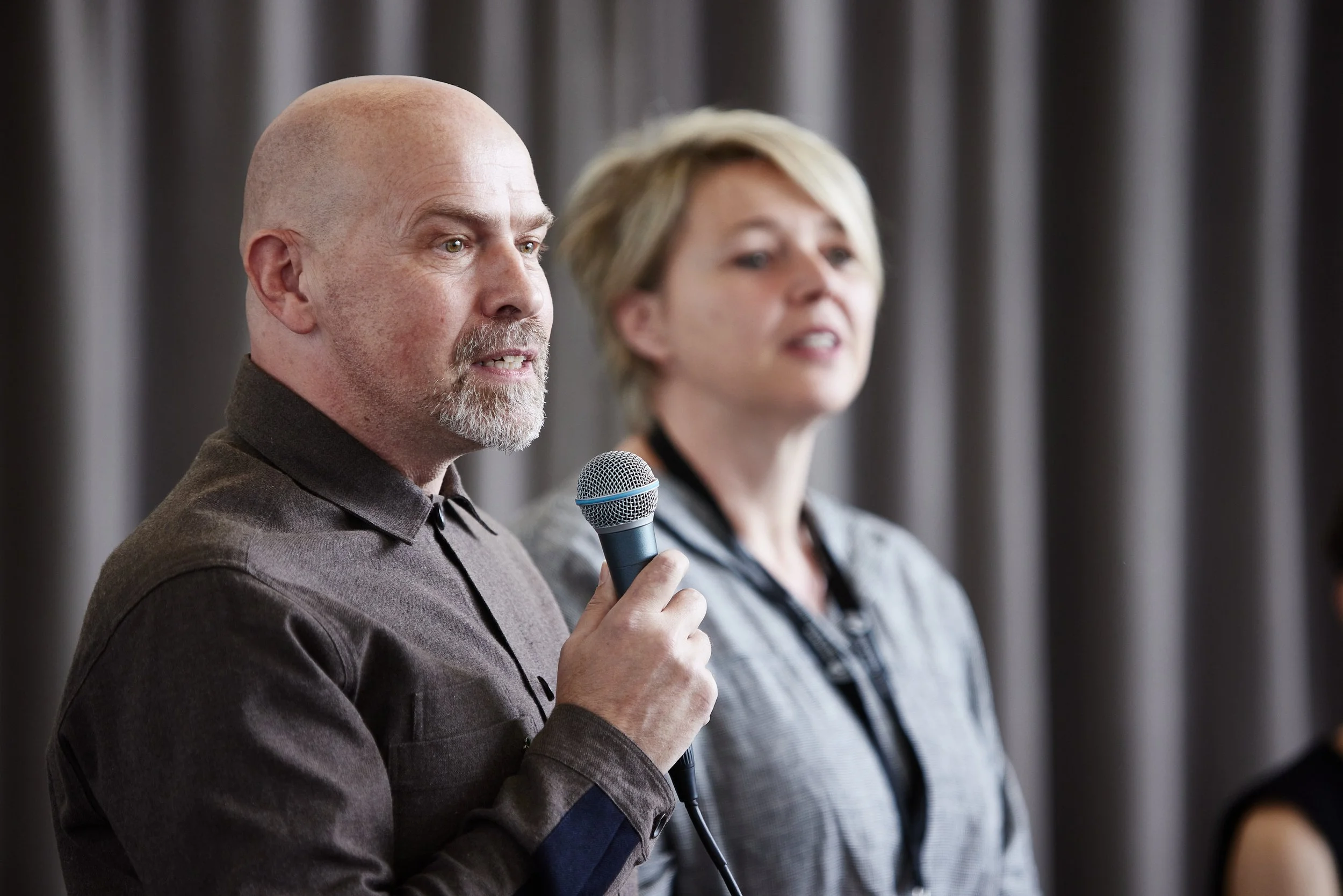A man holding a microphone and speaking in front of a grey curtain, with a woman standing beside him.