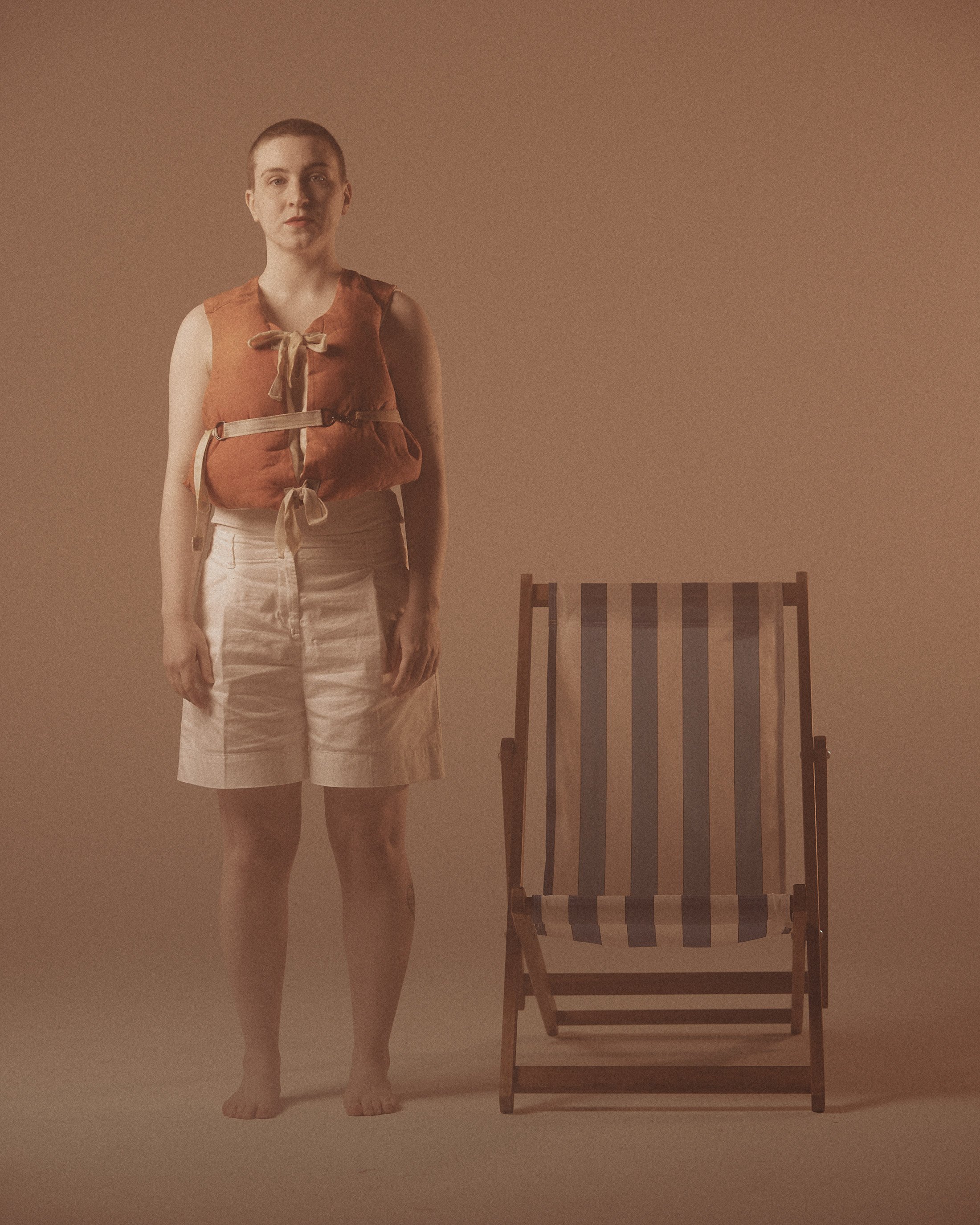 A woman in beige shorts standing next to a wooden lounge chair with blue and beige striped fabric, against a plain beige background.