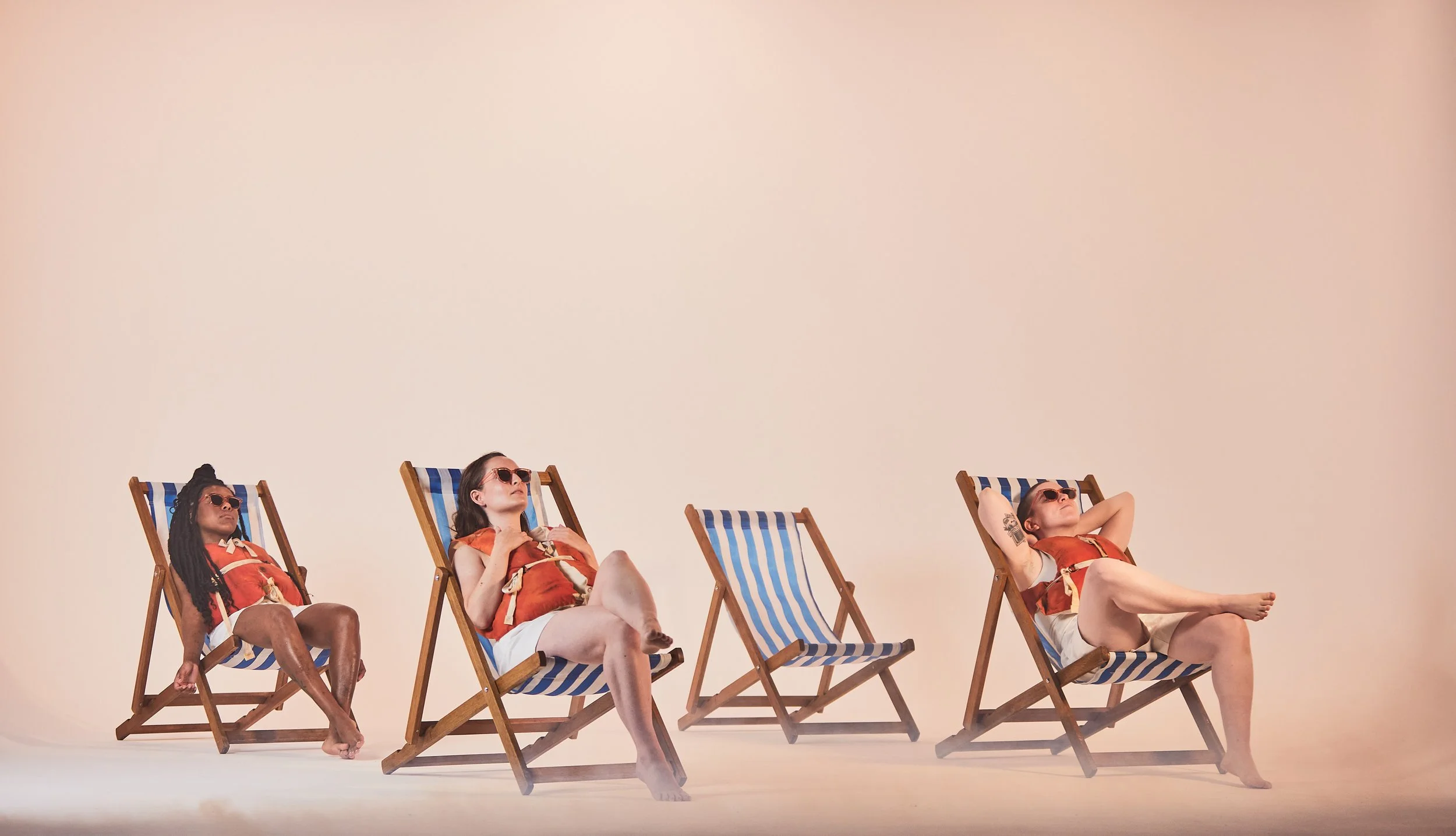 Three women sitting in beach chairs relaxing, wearing sunglasses and red tops, against a plain beige background.