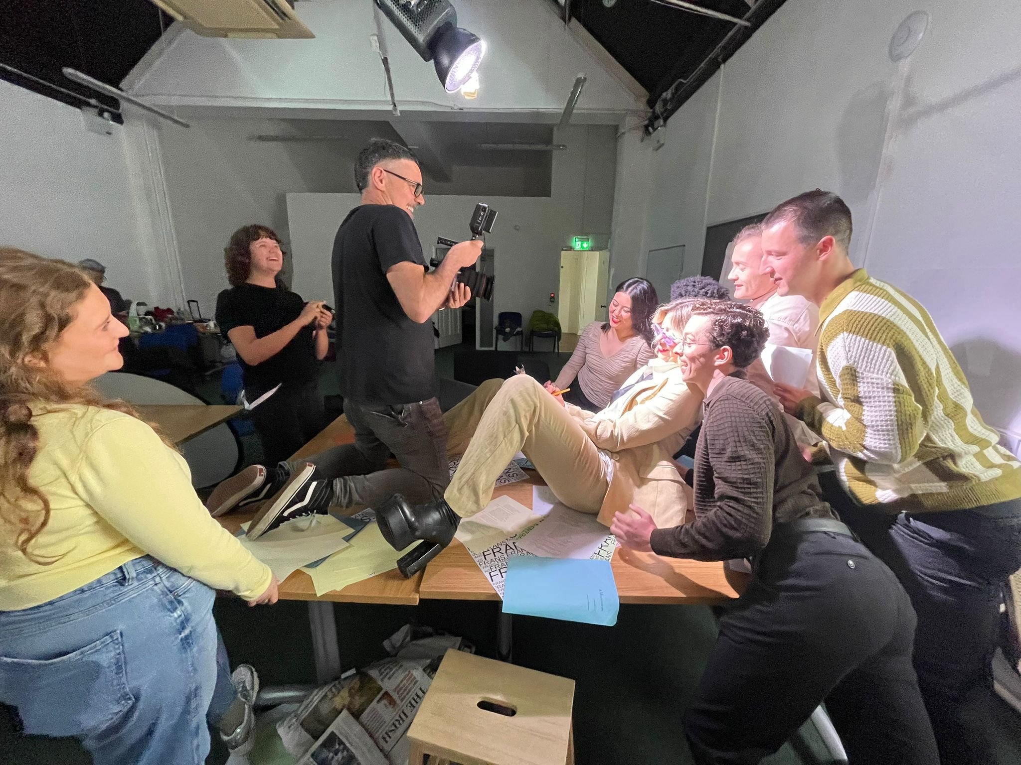 A group of people sitting and standing around a table, with some enjoying a playful moment, while a photographer records the scene.