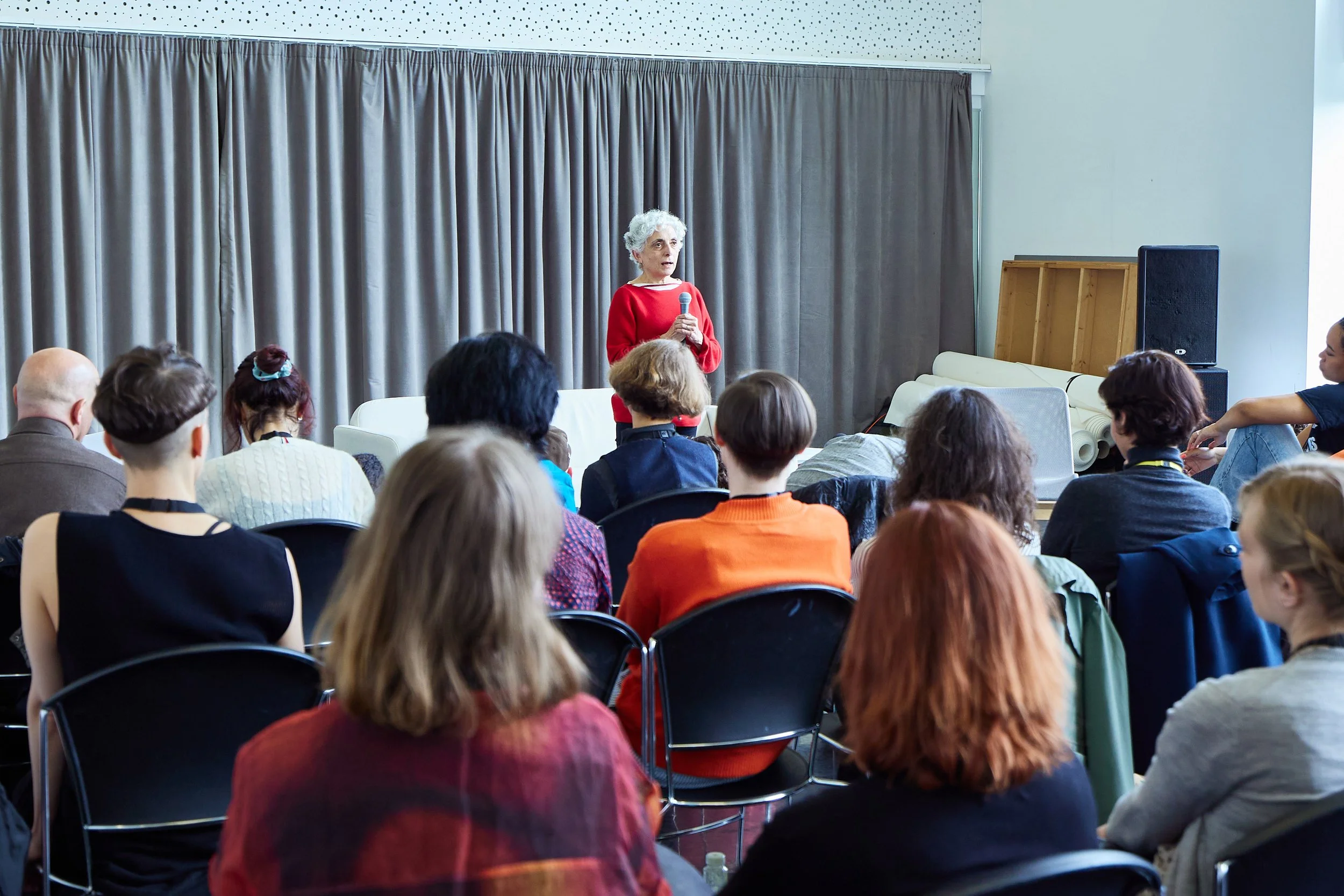 An elderly woman in a red sweater giving a talk to an audience seated in a conference room, with a gray curtain backdrop.