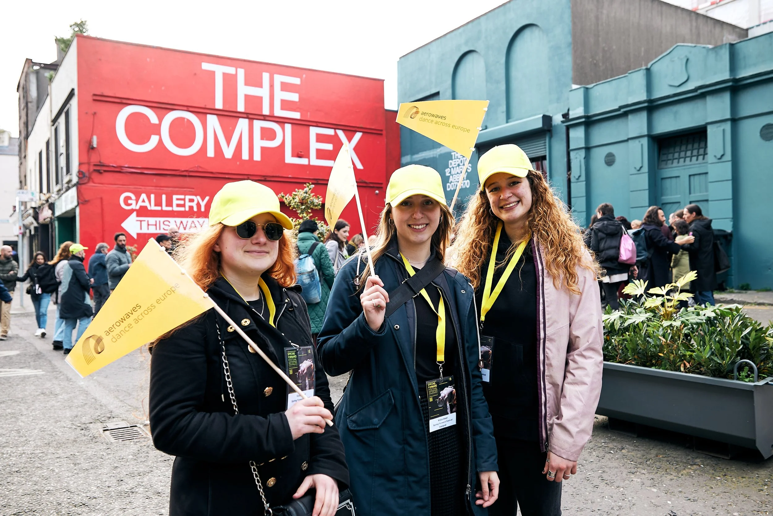 Three women wearing yellow caps and lanyards standing outdoors in front of a building called 'The Complex' with a crowd behind them, holding yellow flags that say 'aerowaves dance across europe'.