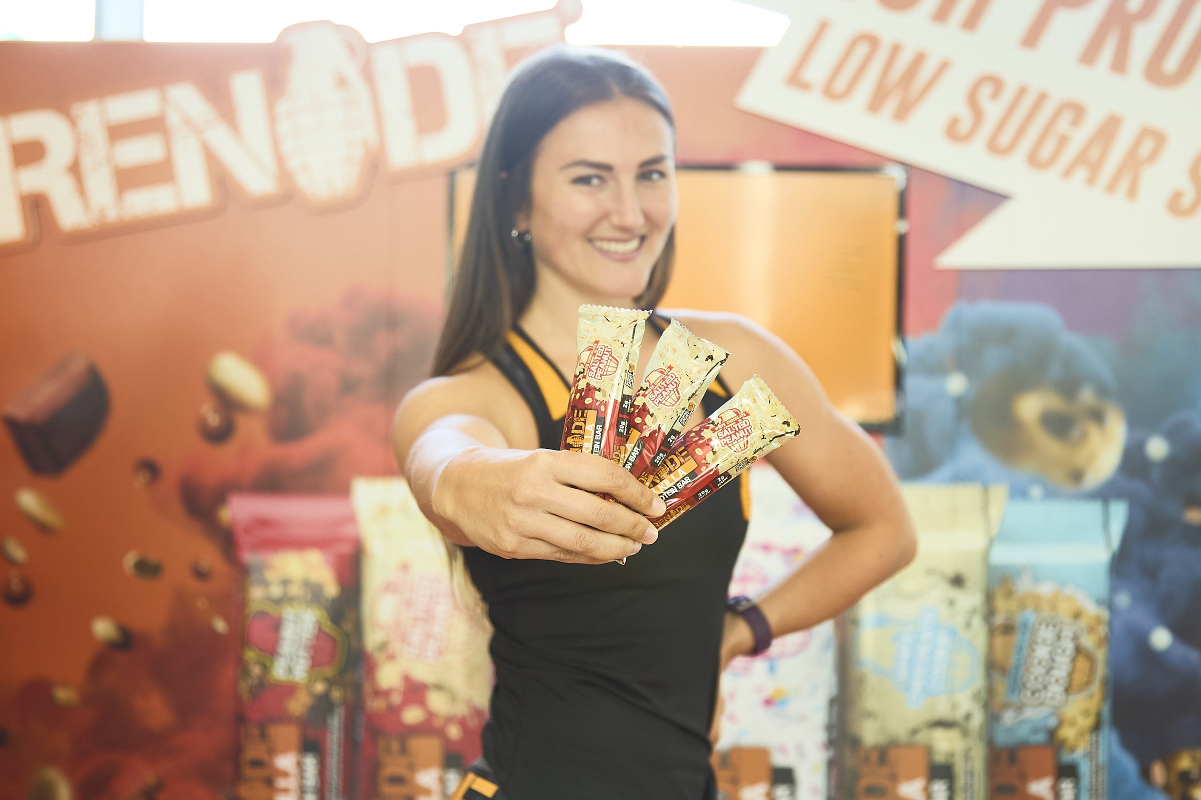 A woman smiling and holding out five salt-flavored protein bars in front of a colorful backdrop with bakery-themed graphics and signs reading 'LOW SUGAR' and 'FATTY PRO'.