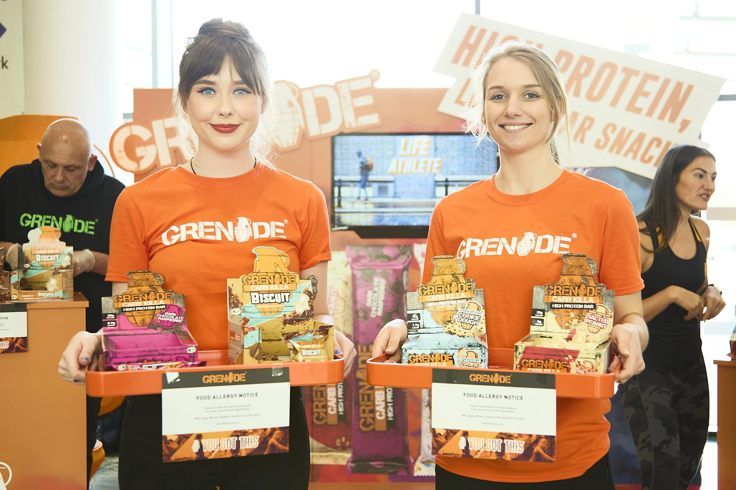 Two women in orange Grenade t-shirts holding orange trays with energy snack bars, at a promotional event with a Grenade display and signs in the background.