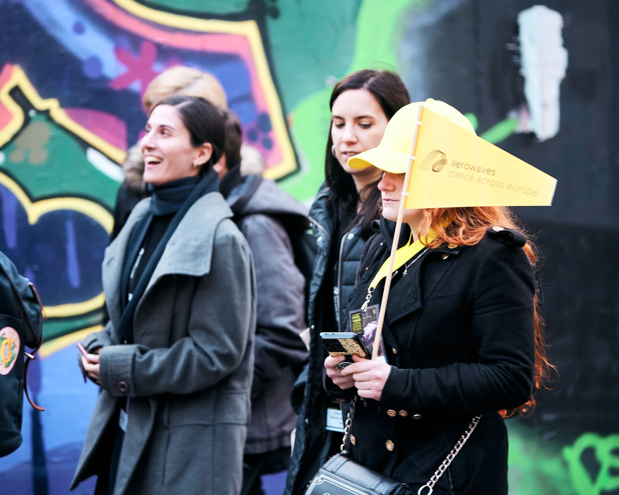 Group of women standing outdoors, some with backpacks, one holding a yellow flag with the text 'aerowaves dance across europe', and others looking at their phones, against a colorful graffiti wall.