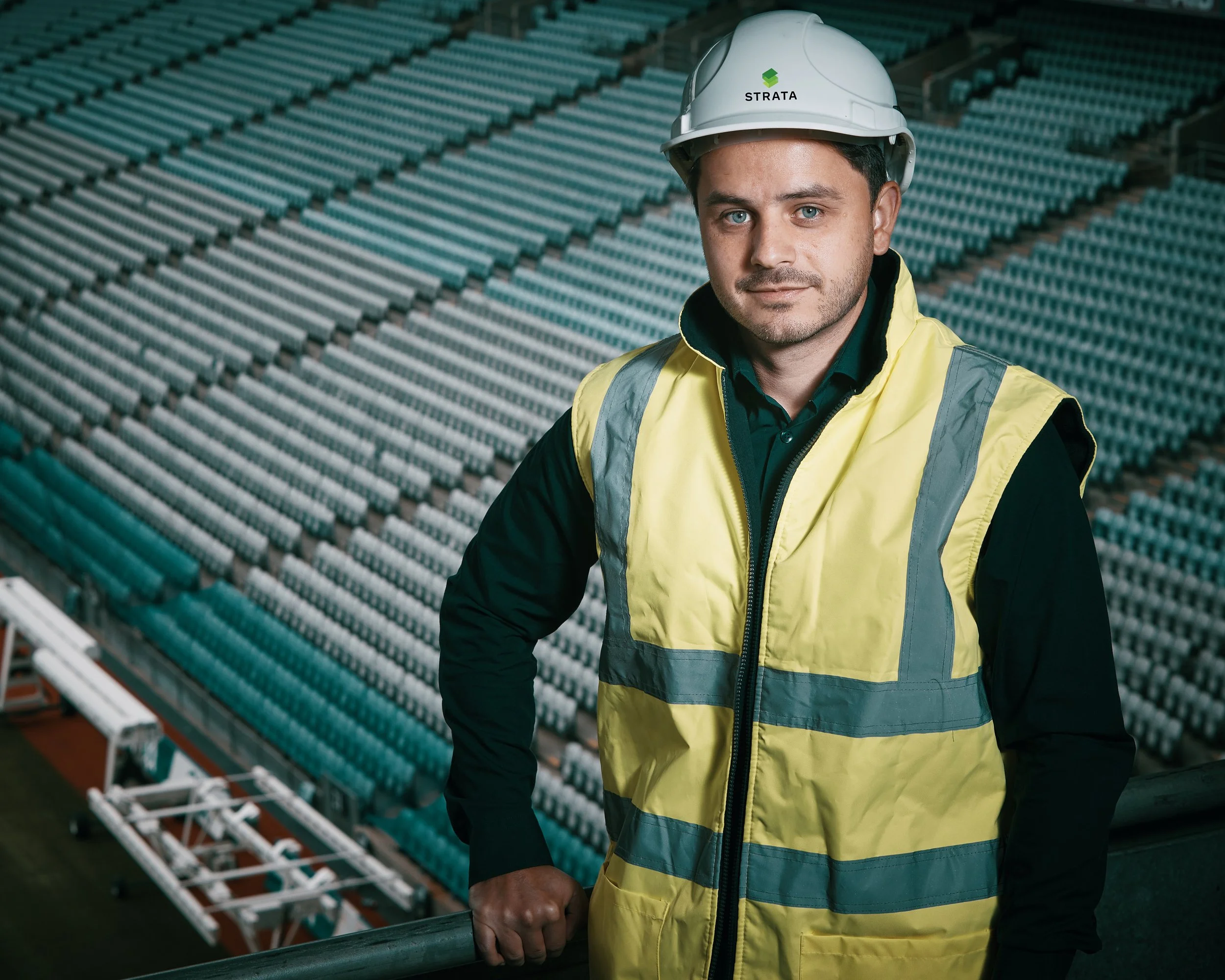 A male engineer wearing a white safety helmet and yellow high-visibility vest stands in front of stadium seating in a professional corporate headshot.