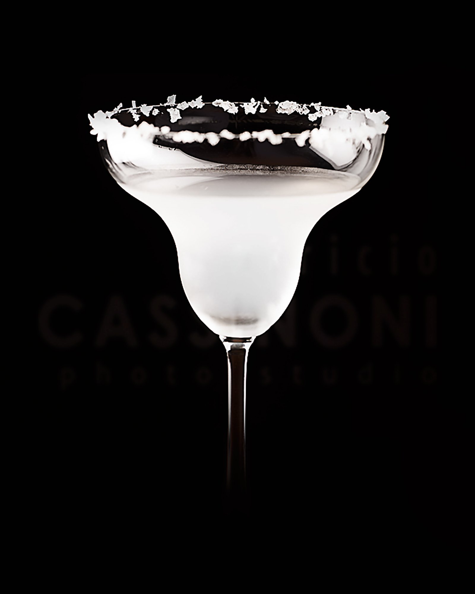 A cocktail glass filled with a white drink, rimmed with salt, against a black background, with a faint logo in the background.
