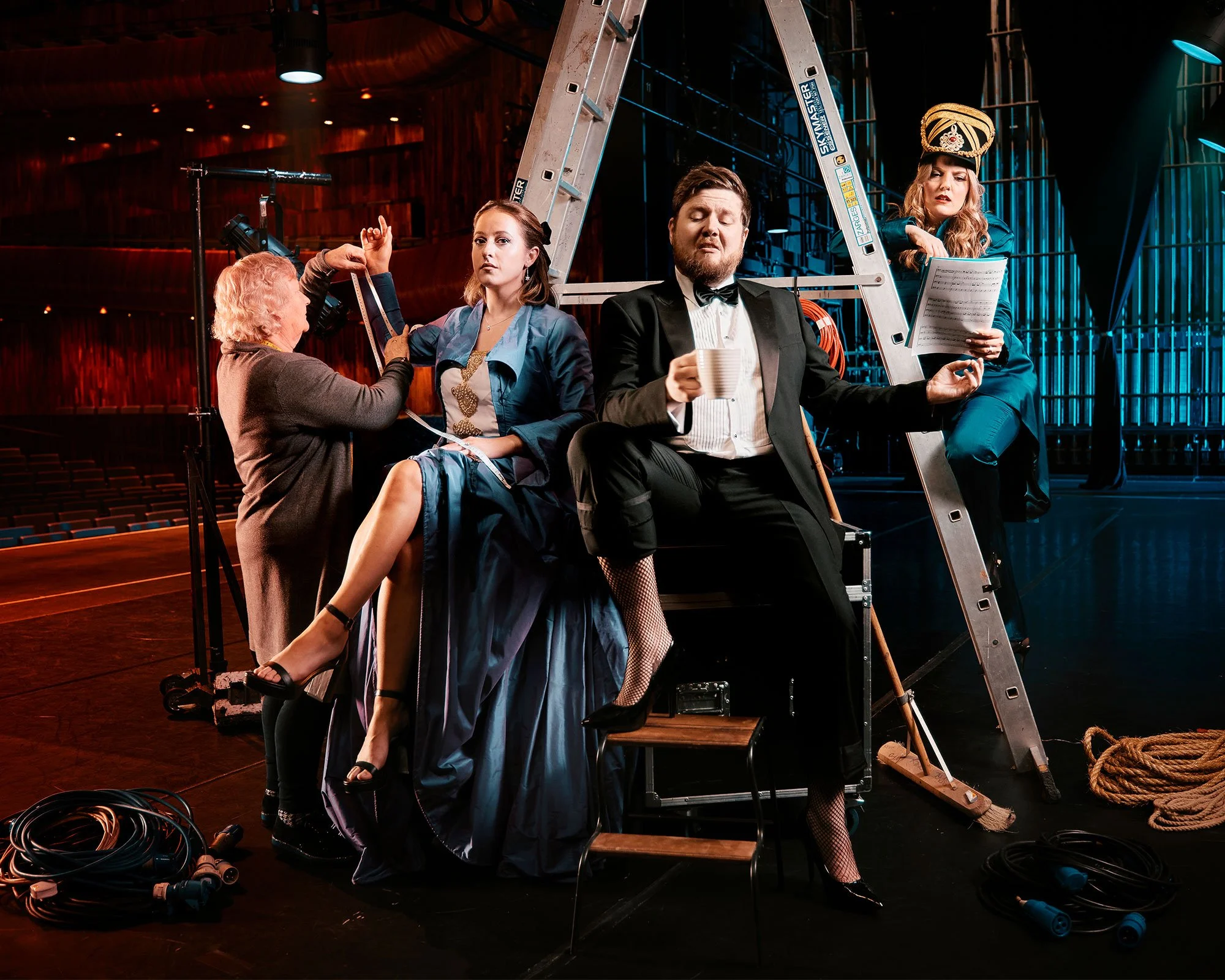 Professional promotional photo of actors in costume during a theatrical rehearsal on stage with technical crew and set pieces shooting in location.