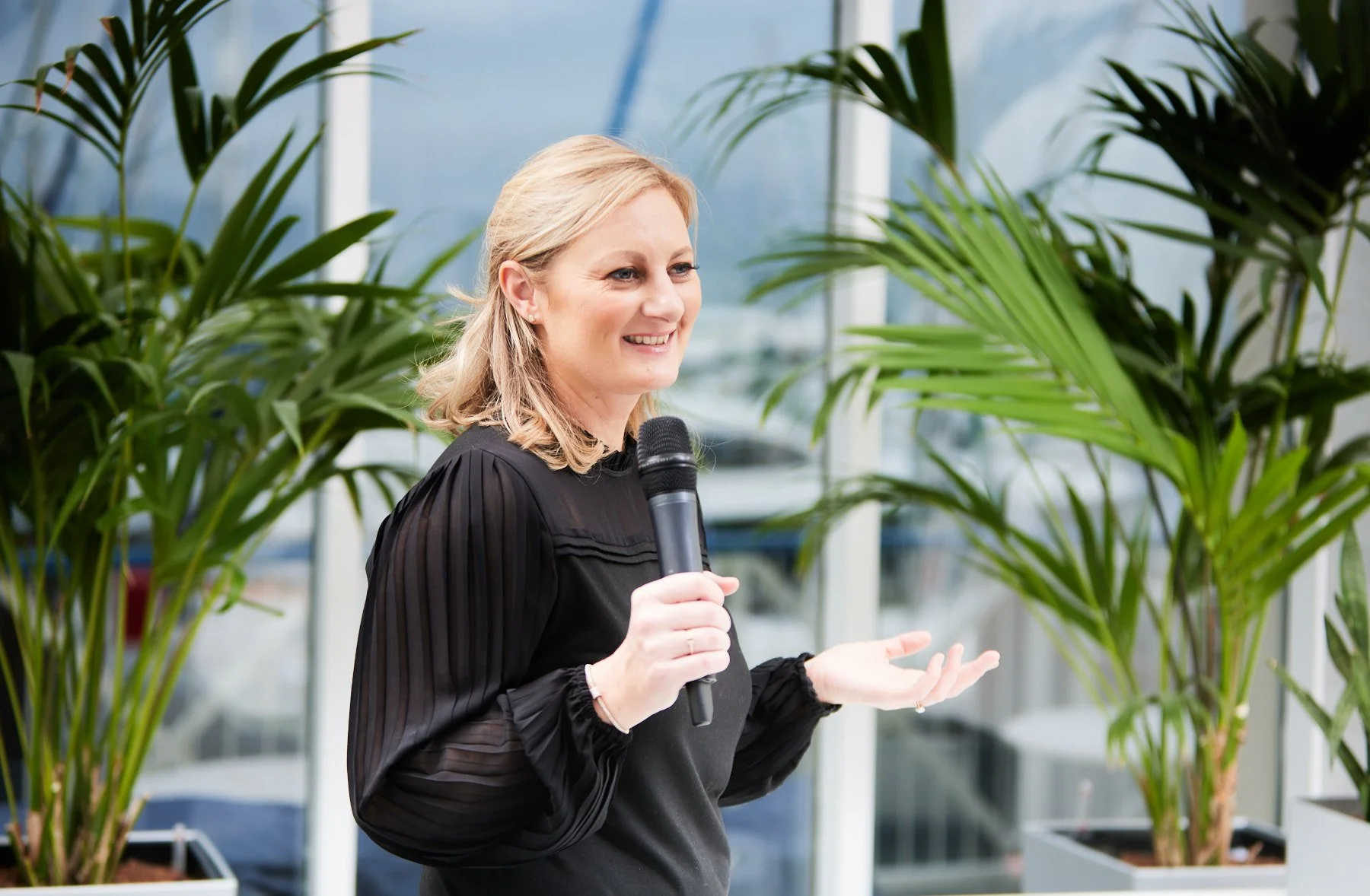 Corporate image of a woman with blonde hair smiling while speaking into a microphone in an indoor space surrounded by large green plants.