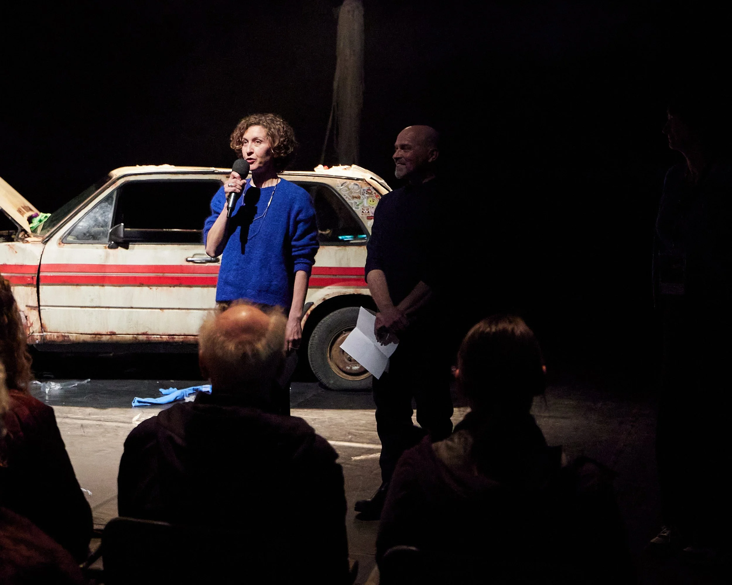 A woman in a blue sweater is speaking into a microphone on stage with three people in the audience. Behind her is an old, rusty car with a red stripe, inside a dark theater or performance space.