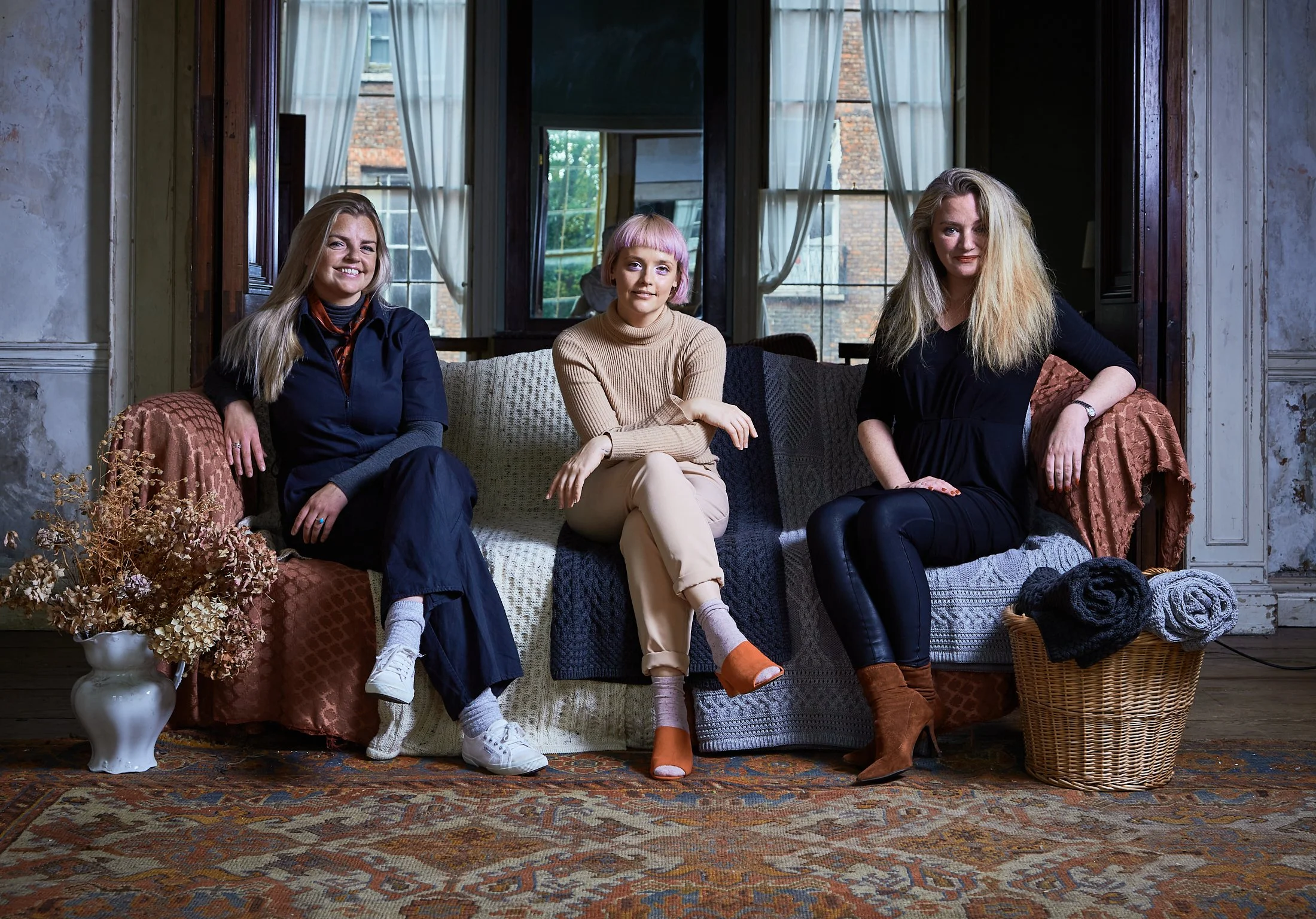 Advertising commercial professional photo of three women sitting on a couch in a cozy room with large windows, curtains, and a patterned rug.