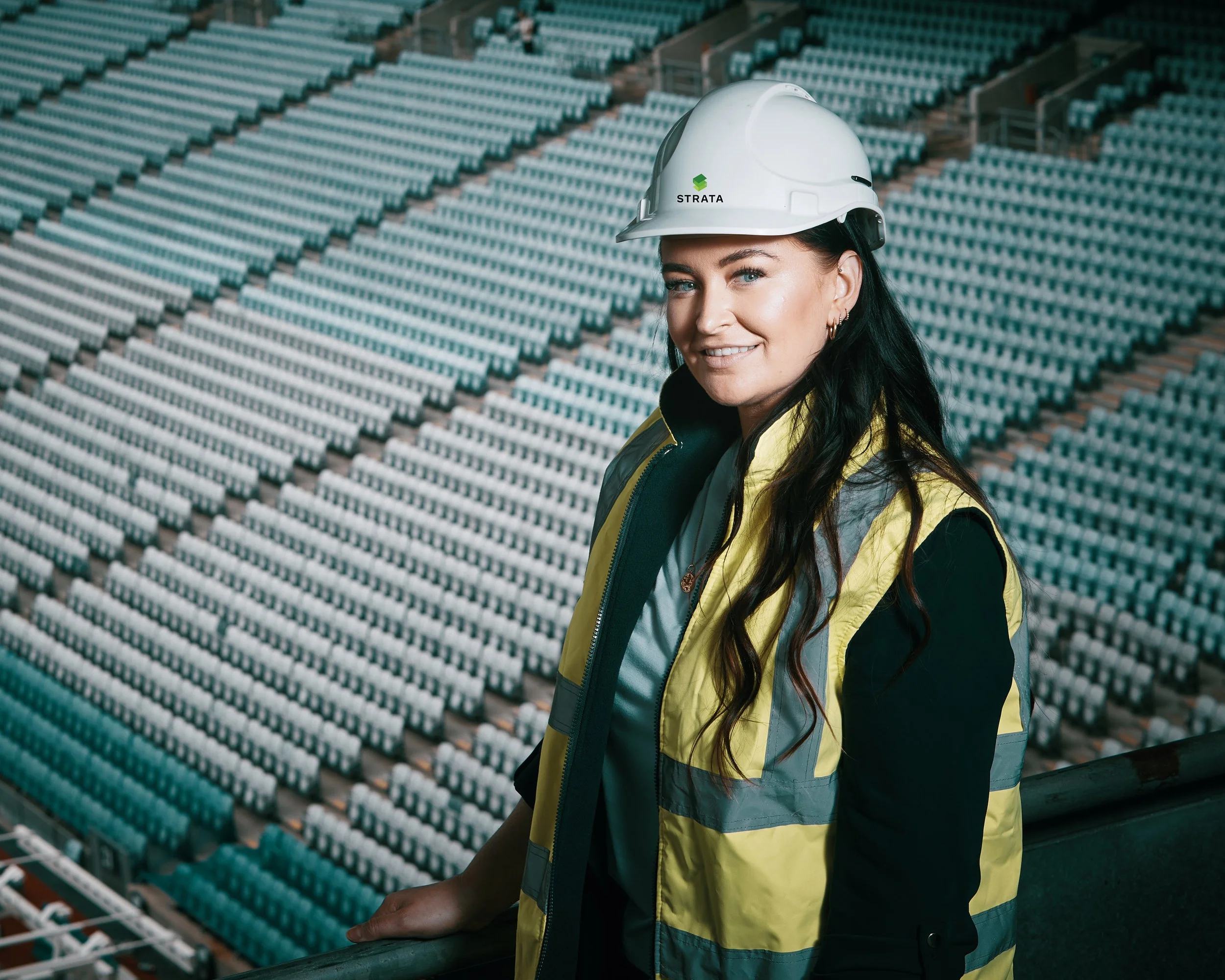 Professional corporate portrait of a woman wearing a white safety helmet and yellow reflective vest standing in a stadium with empty seats.