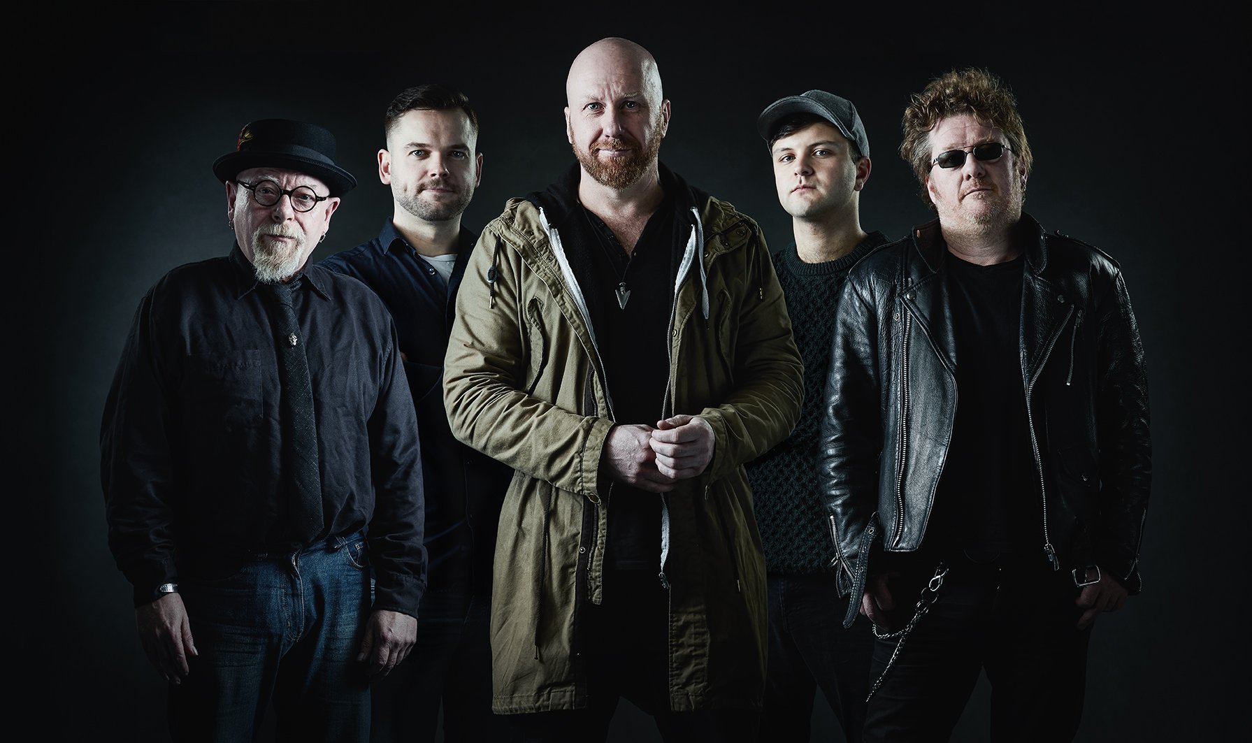Photo in studio of a group of five men standing together in a dark studio, dressed in casual and leather jackets, with a serious expression.