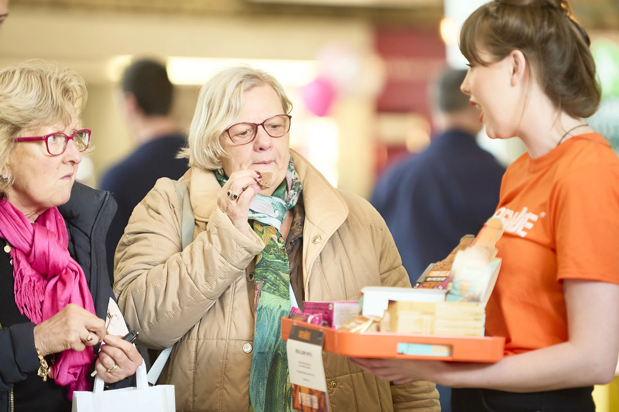 An elderly woman in a beige coat tasting a snack from a tray held by a young woman in an orange shirt. Another woman with pink glasses and a pink scarf watches nearby, in a busy indoor setting.