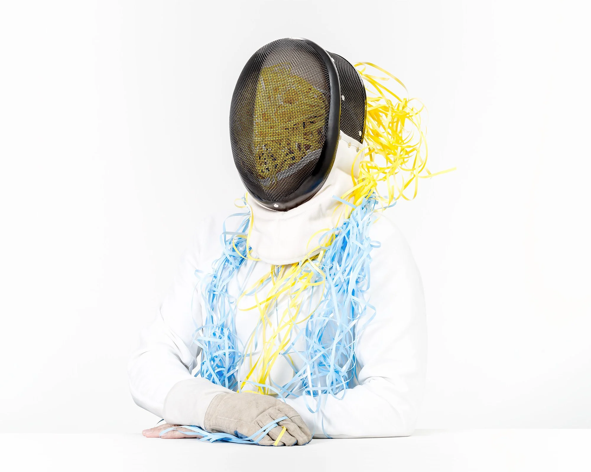 Person wearing a fencing mask with yellow and blue streamers hanging from it, white protective clothing, and a glove, sitting at a white table against a plain white background.