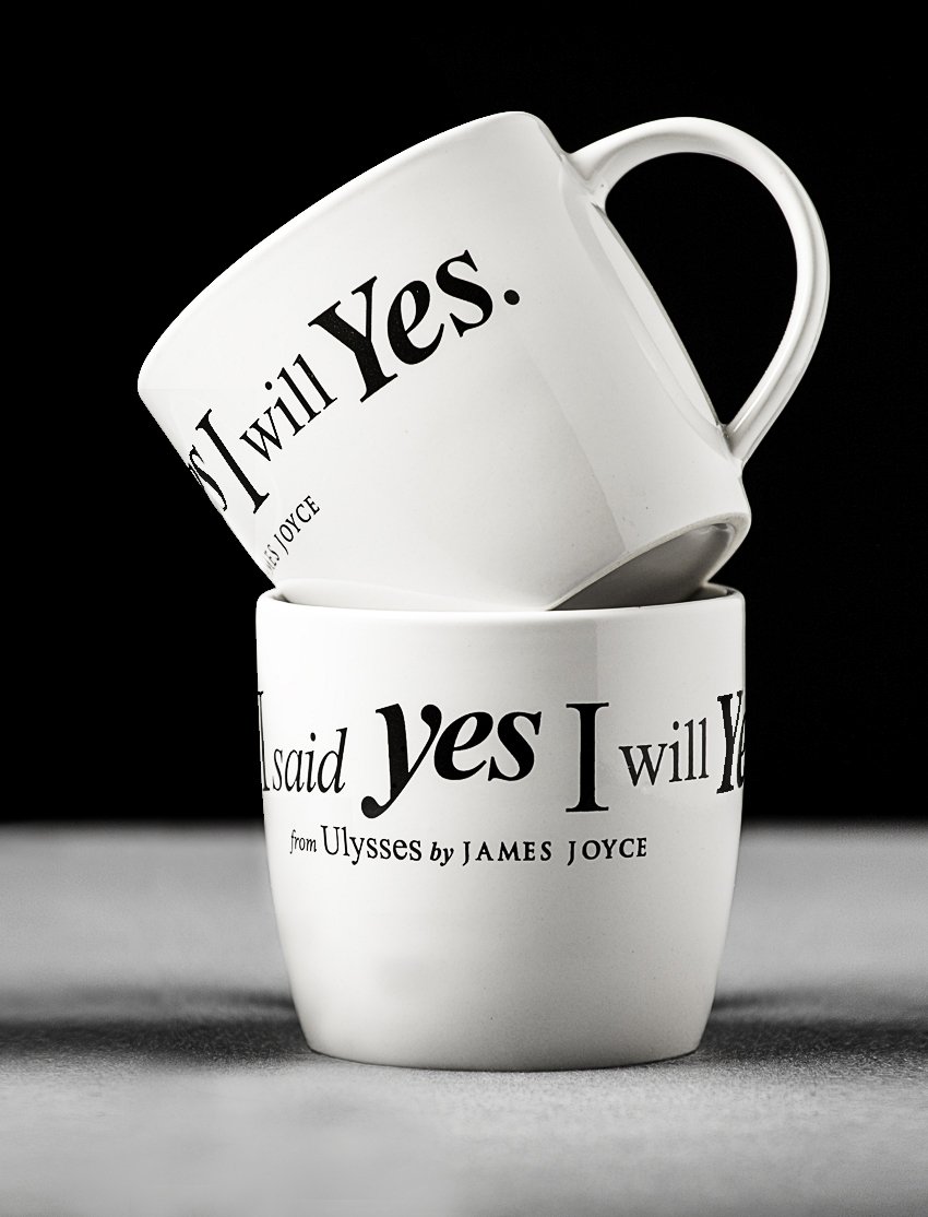 High end advertising profession product photo taken in studio of a two white ceramic mugs with the quote from James Joyce's Ulysses, "Yes. I will Yes." stacked on top of each other, with the top mug tilted.