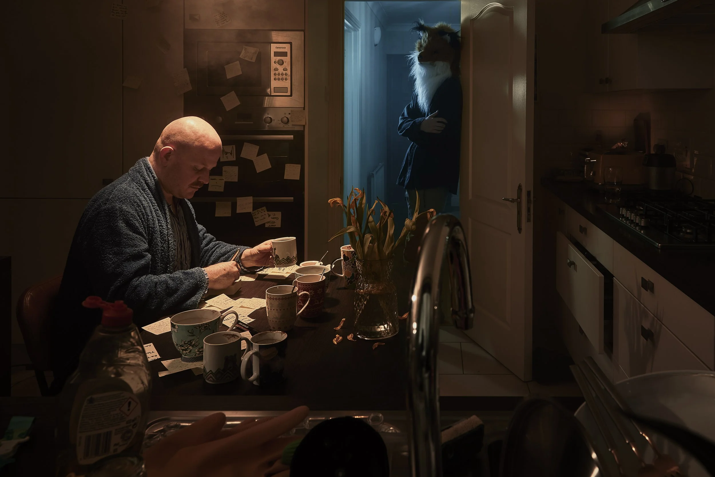A man sitting at a kitchen table with multiple coffee mugs, reviewing notes, while a person wearing a fox mask stands in a doorway, illuminated by blue light, in a dimly lit kitchen.