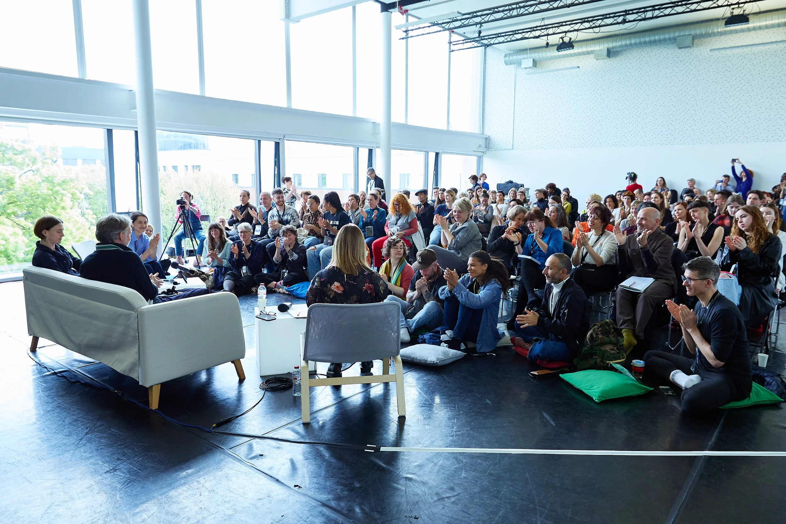 A diverse group of people attending a panel discussion or presentation in a bright, spacious room with large windows and high ceilings. Some are sitting on cushions on the floor, others on chairs, applauding the speakers at the front.