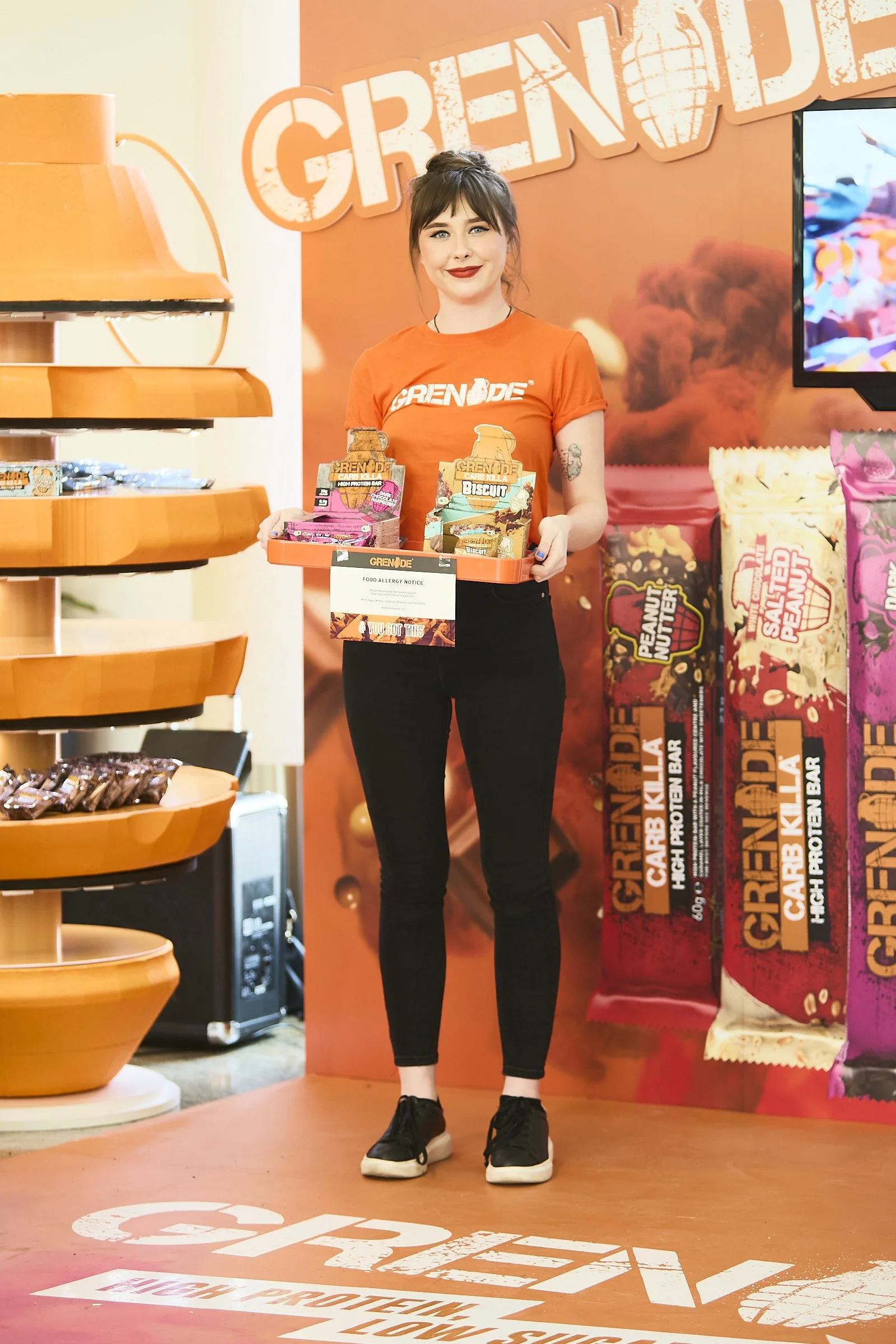 A woman in an orange t-shirt and black pants holds a tray of Grenade brand snack bars at a promotional event with large snack packages in the background.