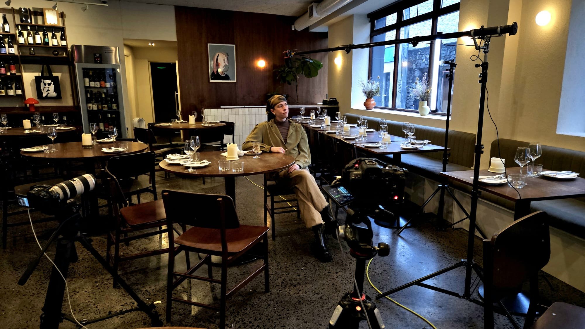A woman sitting at a round dining table in a restaurant or cafe, with filming equipment around her, including a camera on tripod and lighting setup.