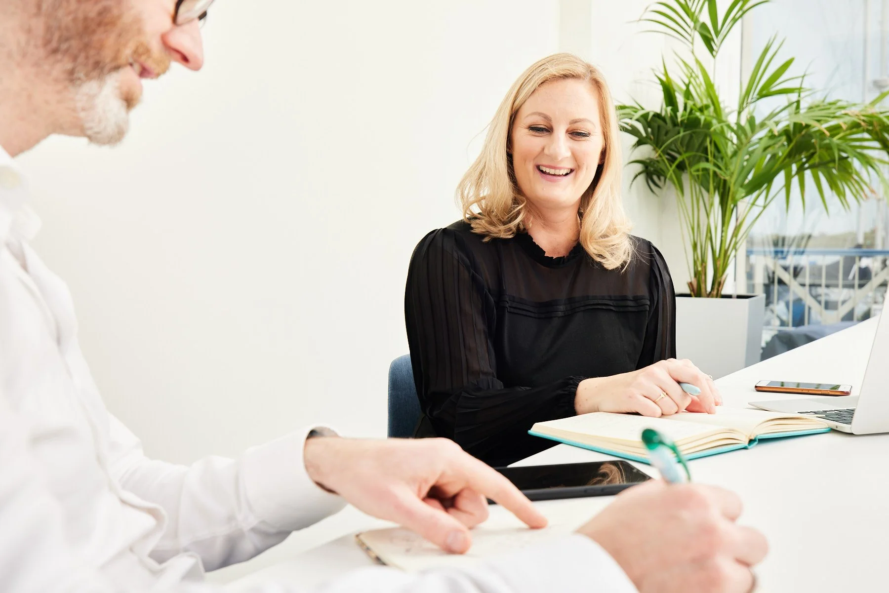 high quality photo of a woman with blonde hair smiling and sitting at a desk with a notebook and open laptop, engaging in a conversation with a man writing in a notepad during a business meeting in a corporate environment in location