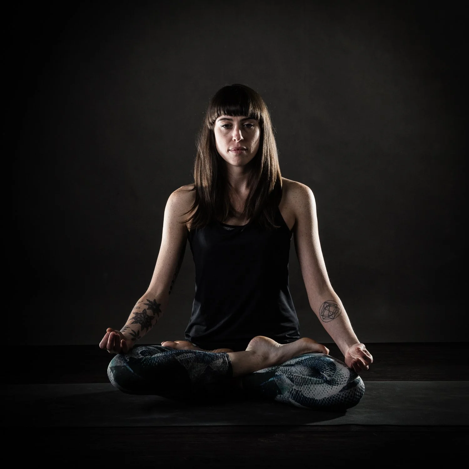 A woman in a black tank top and patterned leggings sitting cross-legged in a meditative pose in a dark space.
