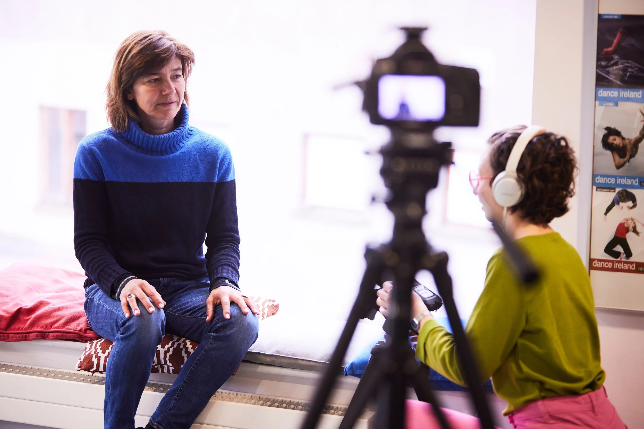 A woman being interviewed on a bed, with a camera and a woman with headphones recording her in a bright room with posters on the wall.
