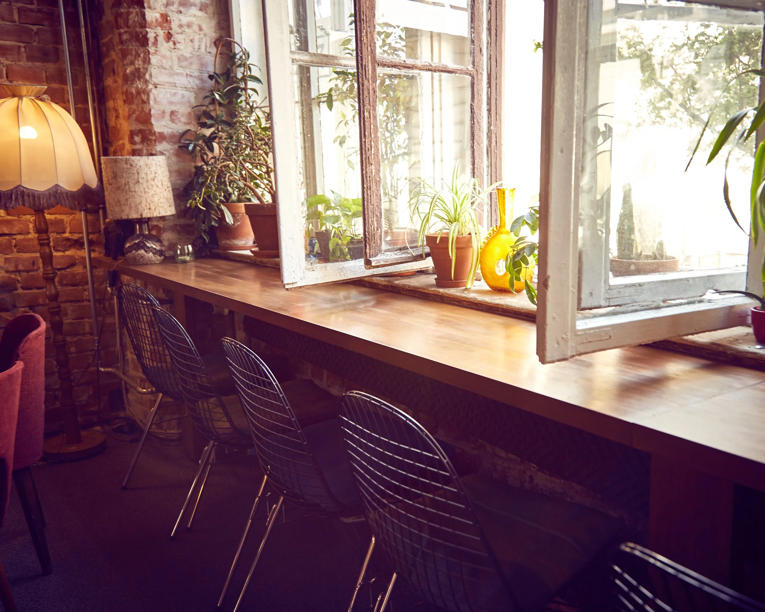 Bright interior of a cozy cafe or home with a large wooden windowsill decorated with potted plants, sunlight streaming in, and a vintage brick wall on the left, accompanied by black wire chairs and a standing lamp.
