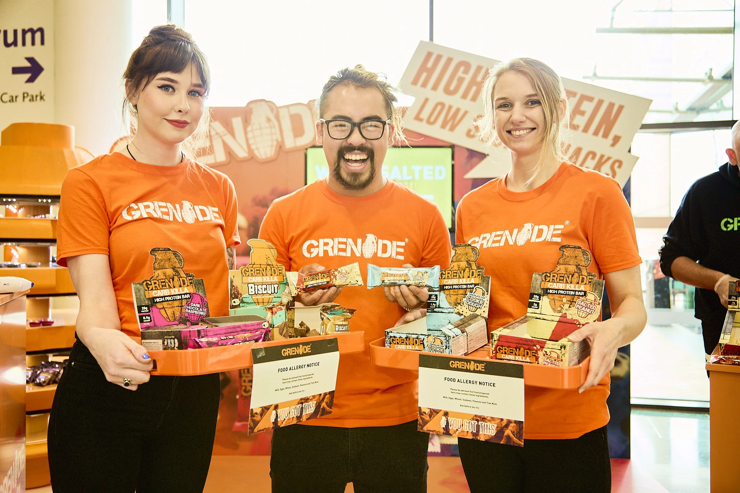 Three people wearing orange T-shirts with 'GRENDE' logo, standing behind orange trays filled with protein snack bars, smiling at the camera inside a store with signs and shelves in the background.