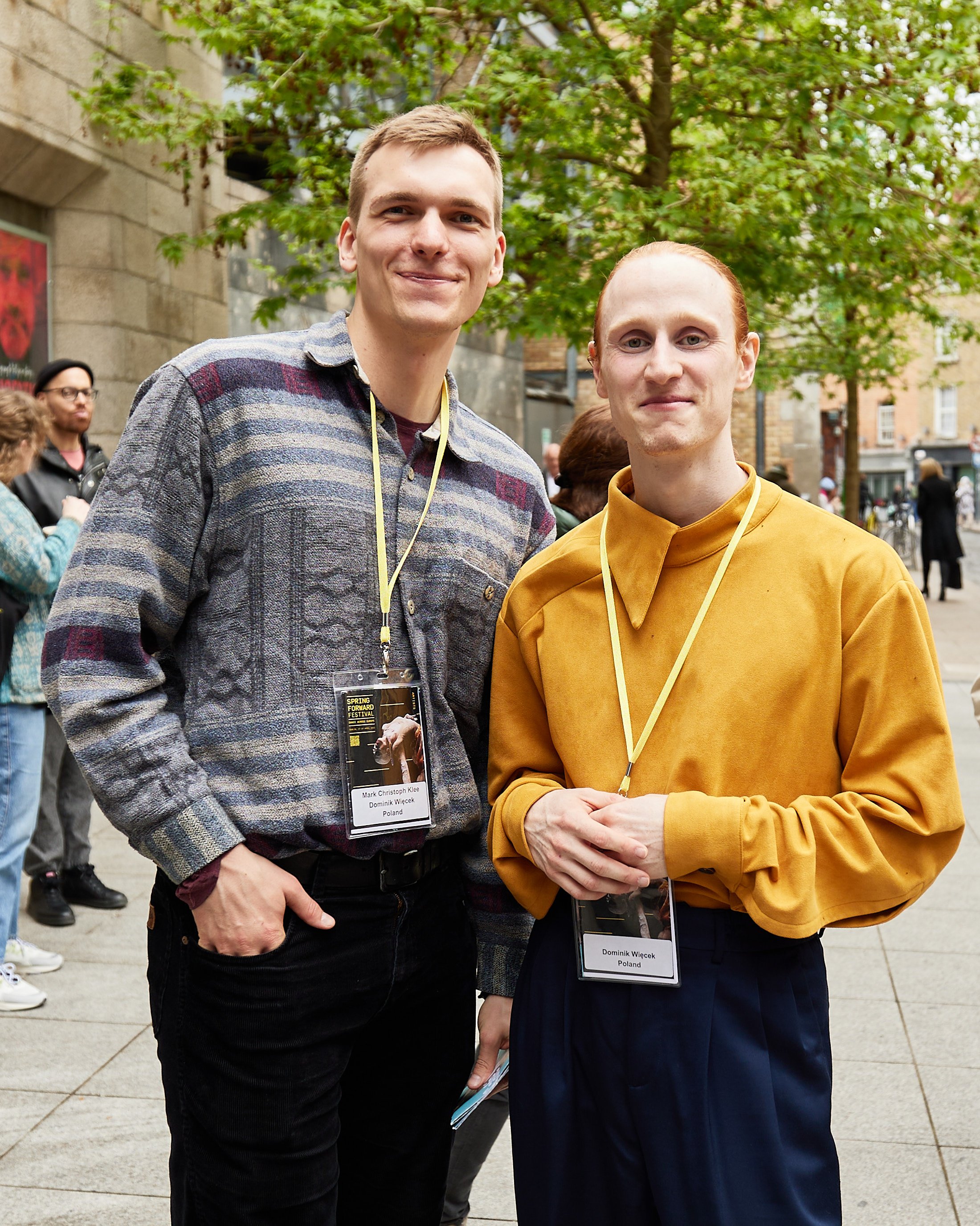 Two young men standing outdoors with trees and several people in the background. They are wearing badges around their necks and smiling at the camera.