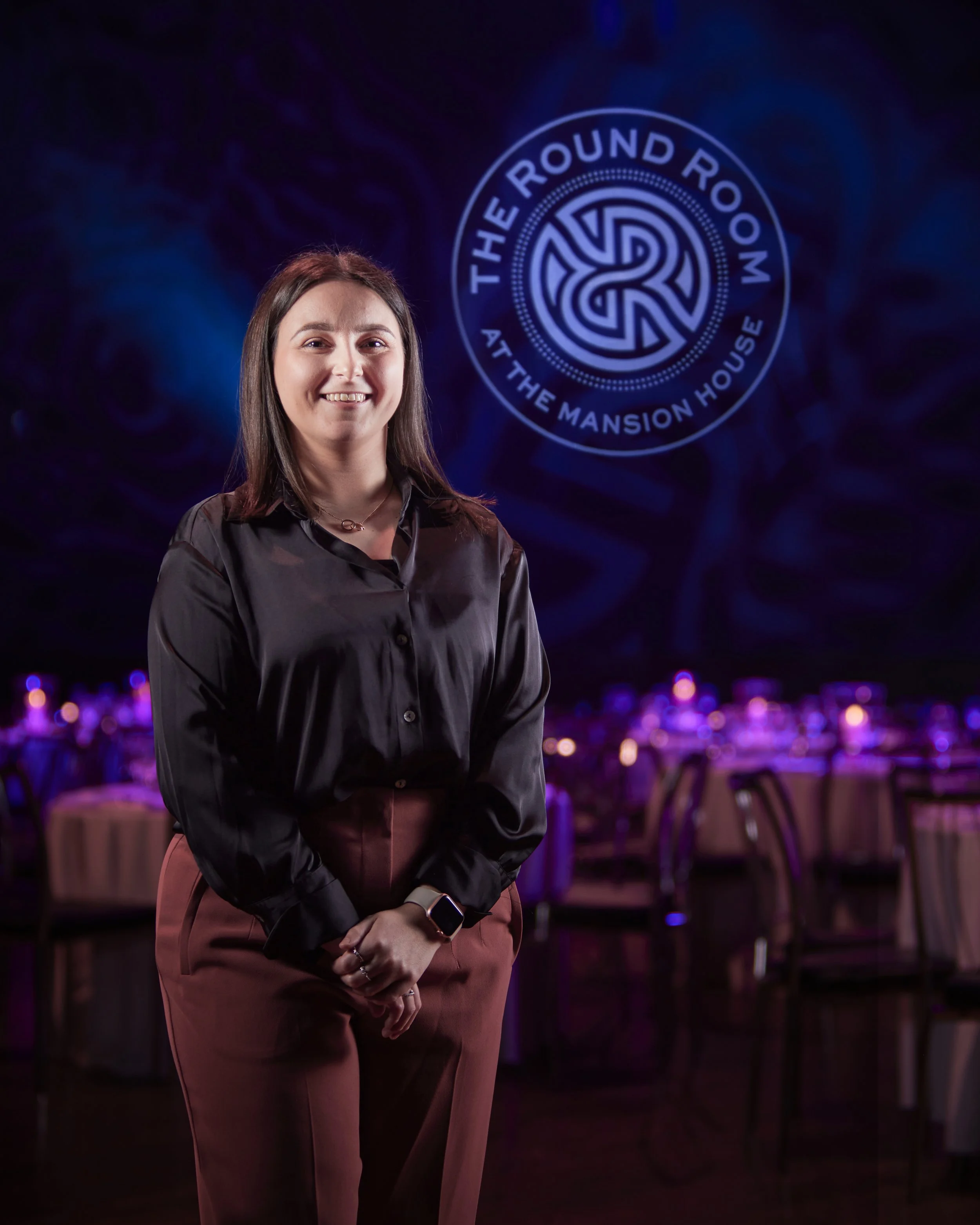 Corporate portrait photography of a woman standing in a dimly lit event space with tables decorated with purple lighting in the background. A circular logo on the wall behind her reads "The Round Room at The Mansion House."