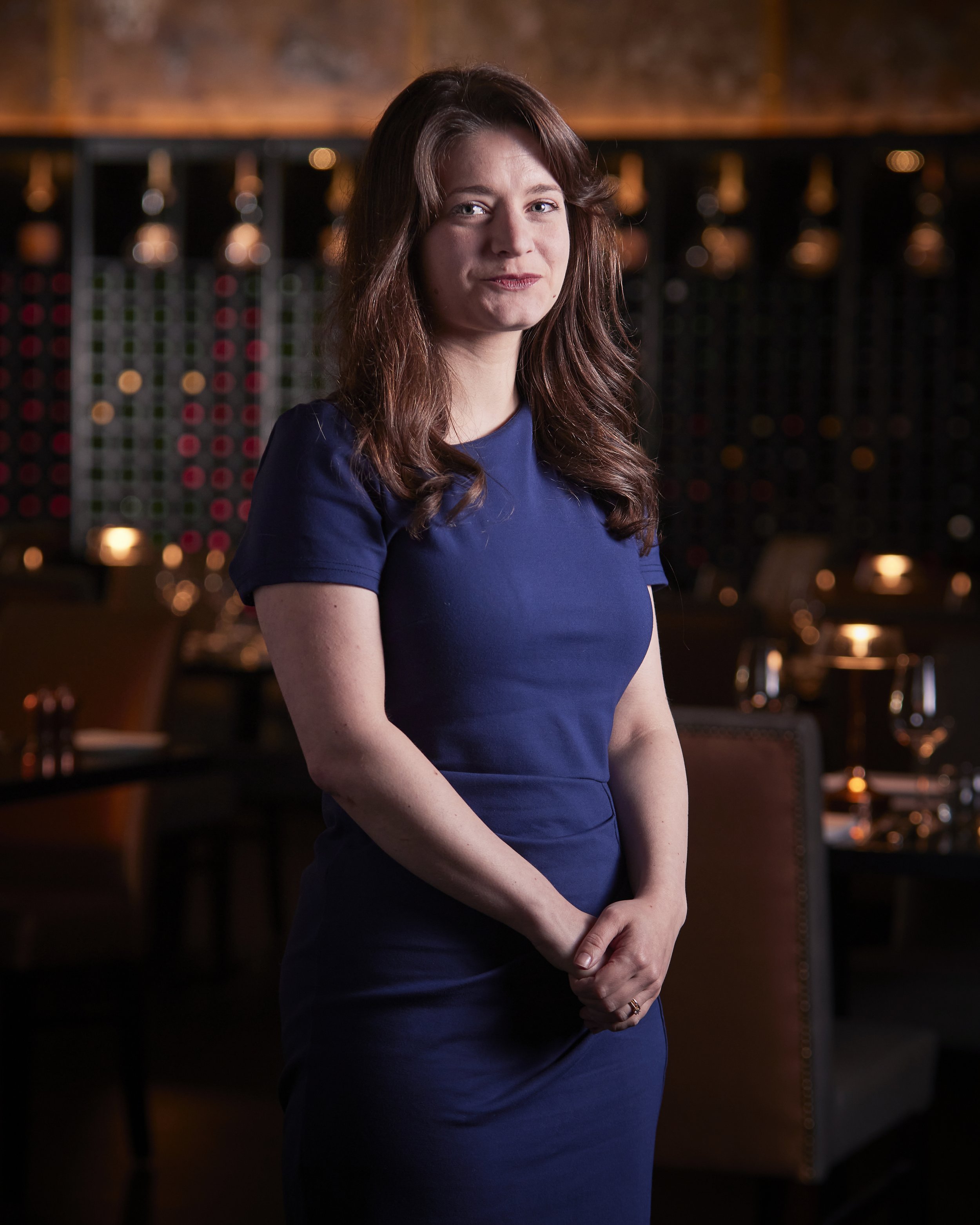 Corporate portrait photography of a Woman with long brown hair wearing a blue dress standing in a dimly lit restaurant or wine bar with wine racks in the background.