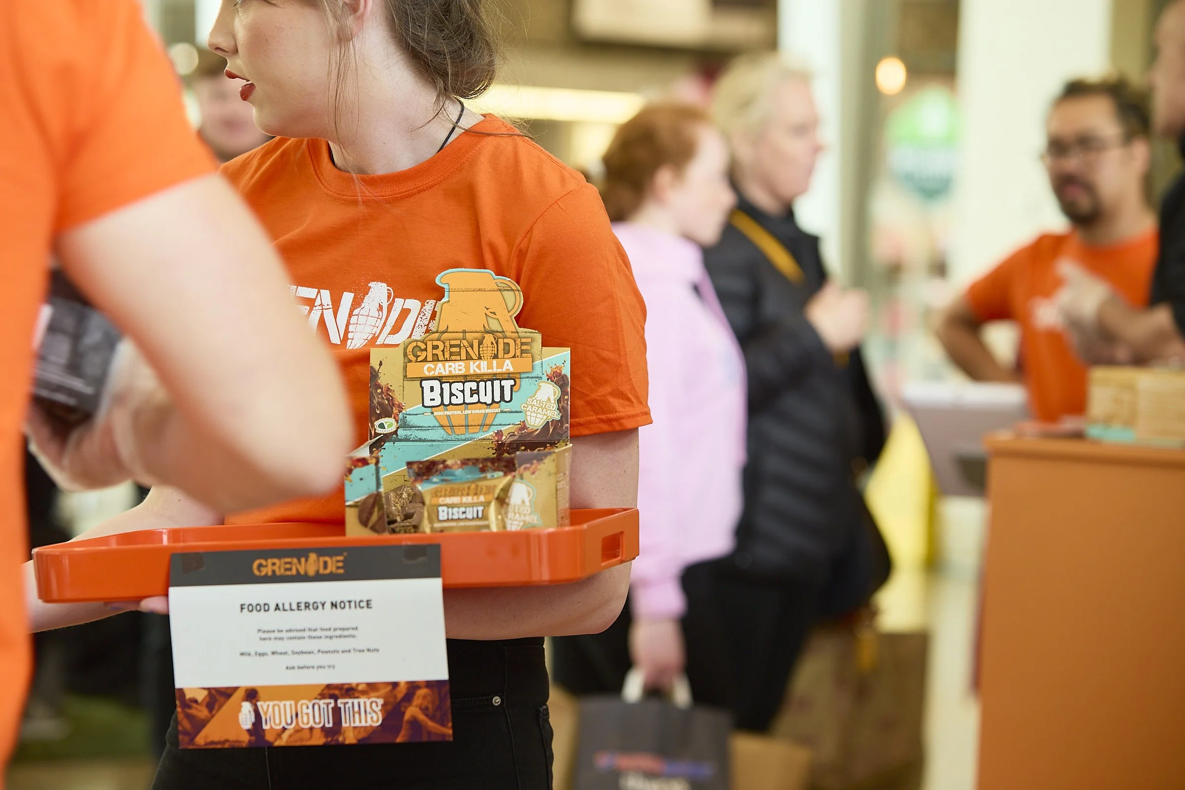 A woman wearing an orange shirt holding a tray with energy protein bars at an indoor event. In the background, several people are standing and talking, some holding bags.