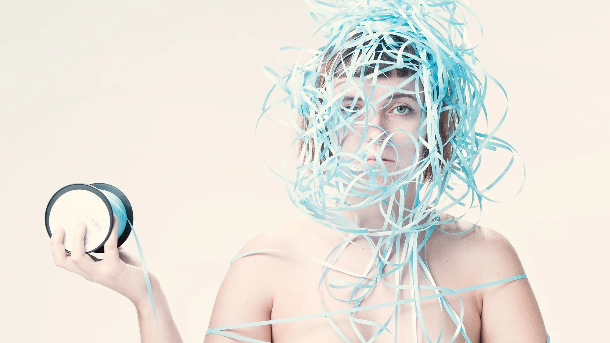 Advertising photo of a woman with blue eyes and short brown hair is partially covered by blue  paper streamers on her head and shoulders. She is holding a spool of blue ribbon in her right hand, shooting in a photo studio with professional lighting.
