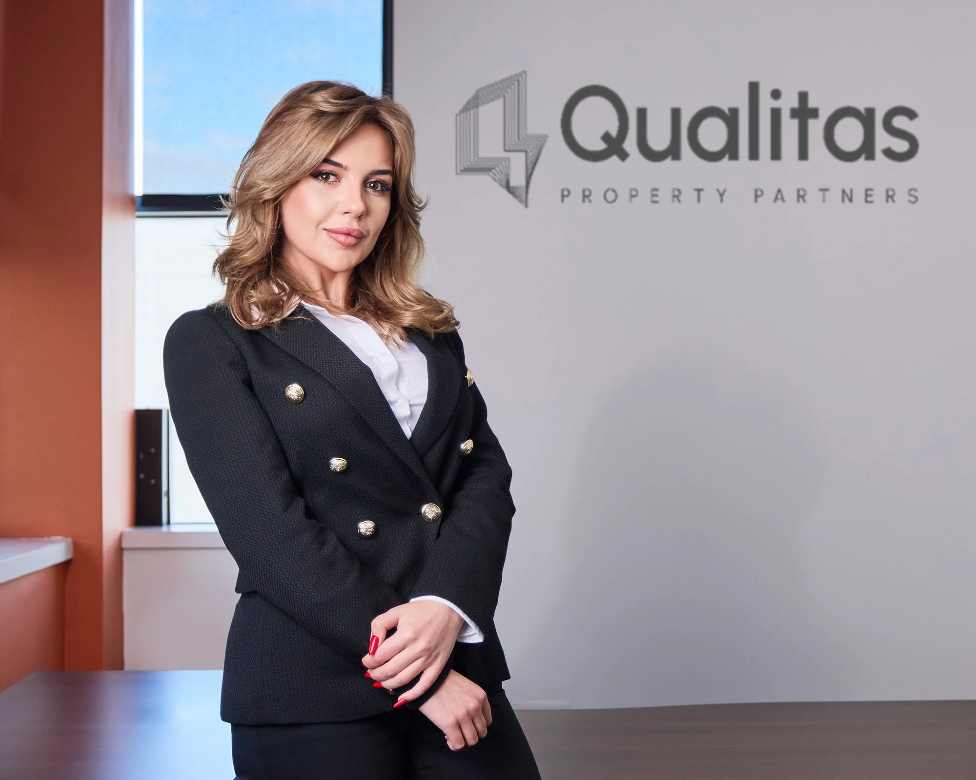 Professional corporate portrait, corporate headshot of a woman with blonde hair wearing a black blazer with gold buttons, white shirt, and red nail polish standing in an office with a logo that reads 'Qualitas Property Partners' on the wall 