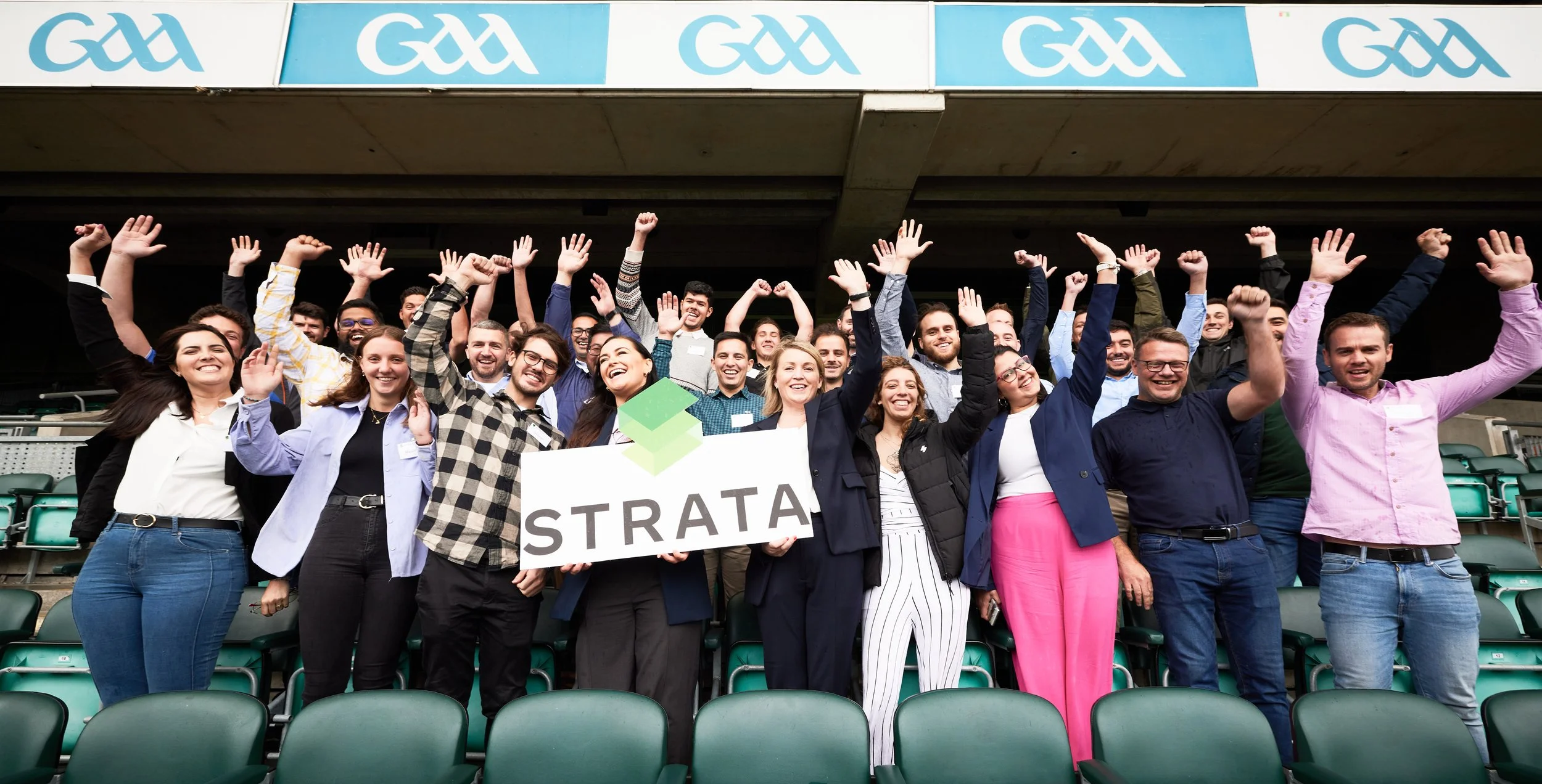 Professional photo of a corporate group of people at a AVIVA stadium holding a sign that says 'STRATA' celebrating, with many raising their hands in the air.