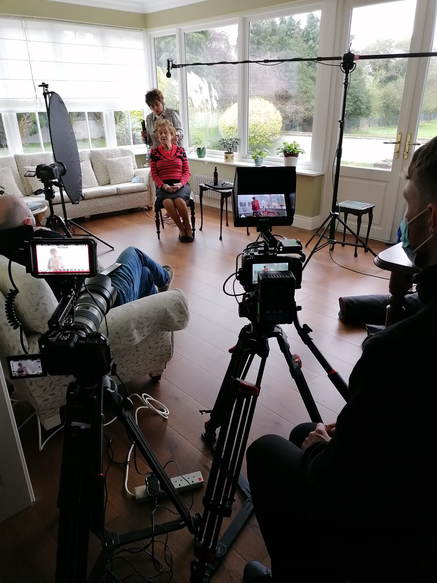 A woman in a red and black striped sweater sitting in a chair in a sunlit room with large windows, surrounded by film equipment including cameras, lights, and monitors, with crew members filming her.