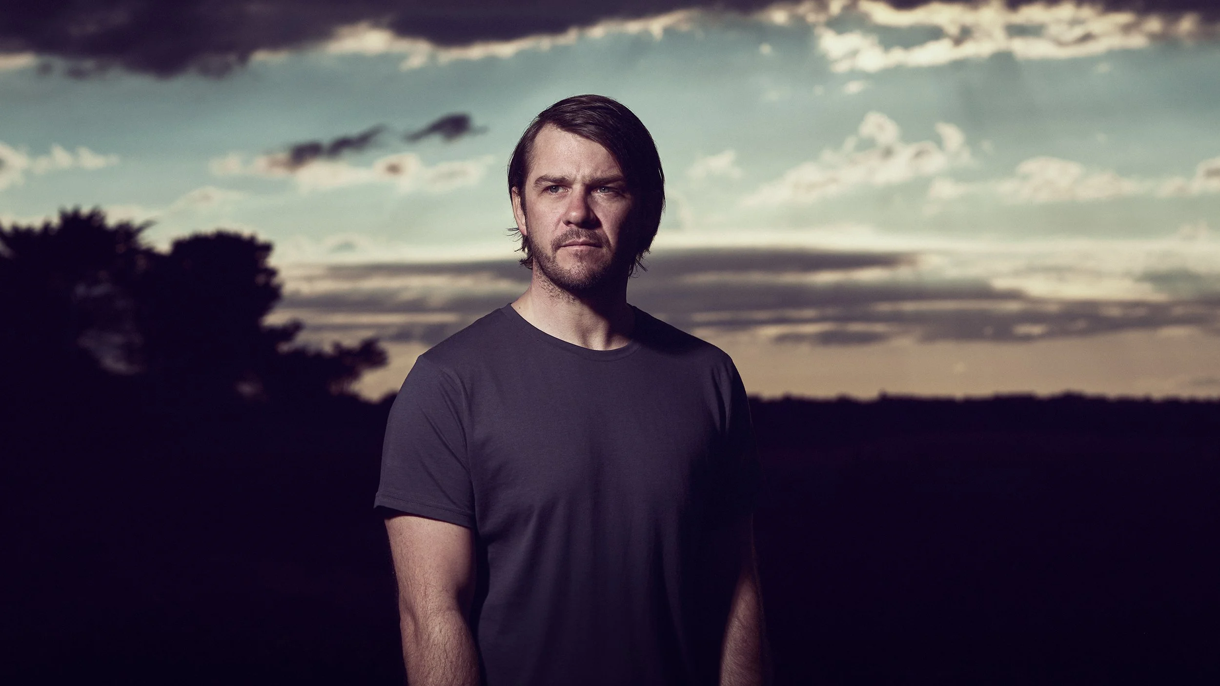 Strong advertising image of man with dark hair and beard wearing a black t-shirt standing outdoors at sunset or dusk, with a cloudy sky and a silhouette of trees in the background, shot in location with a high quality professional lighting.