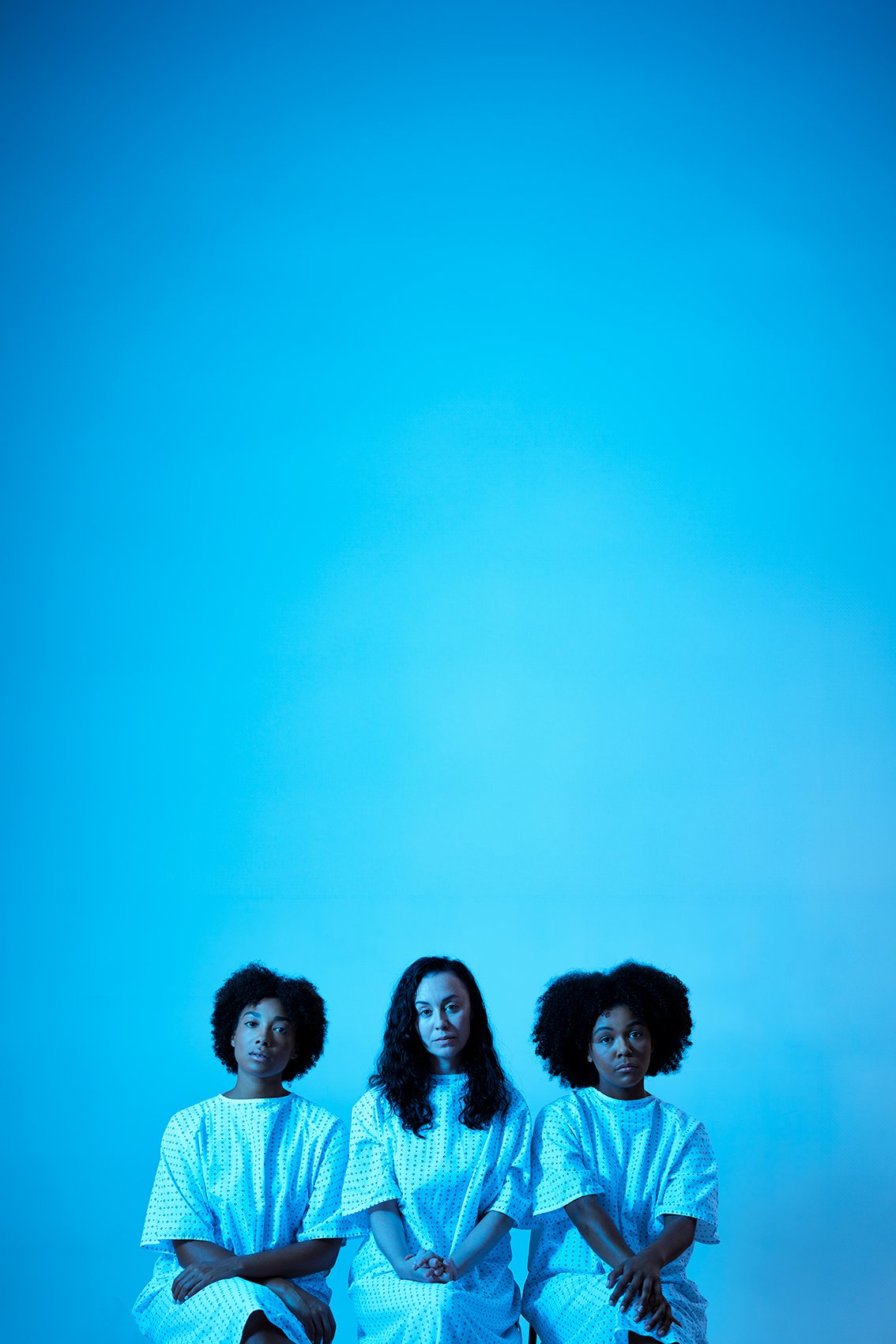 Promotional image of three women sitting in hospital gowns against a blue background in a photo studio with great professional lighting.