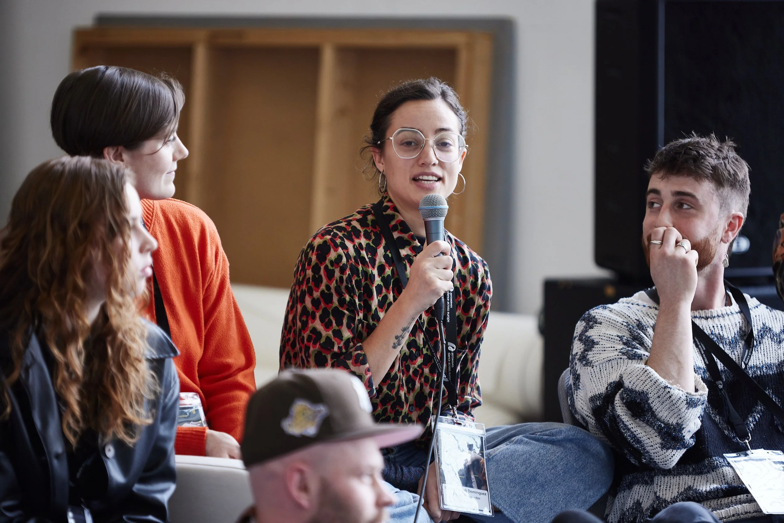 A woman with glasses and hoop earrings, speaking into a microphone at a panel or conference, with other people listening, in an indoor setting.