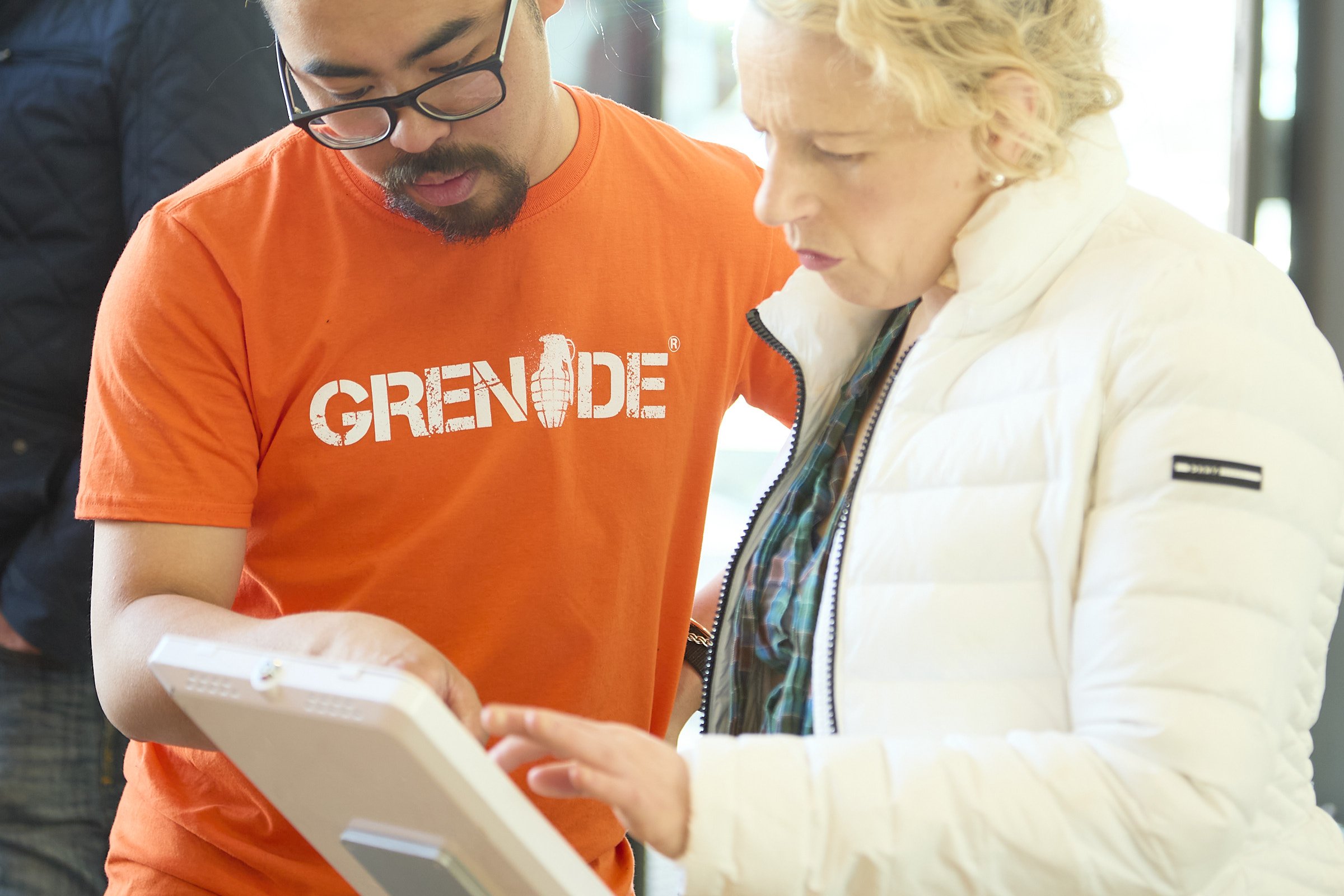 Two people, a man with glasses wearing an orange Grenade shirt and a woman in a white jacket, are looking at a tablet together. They seem to be discussing or reviewing something on the device.