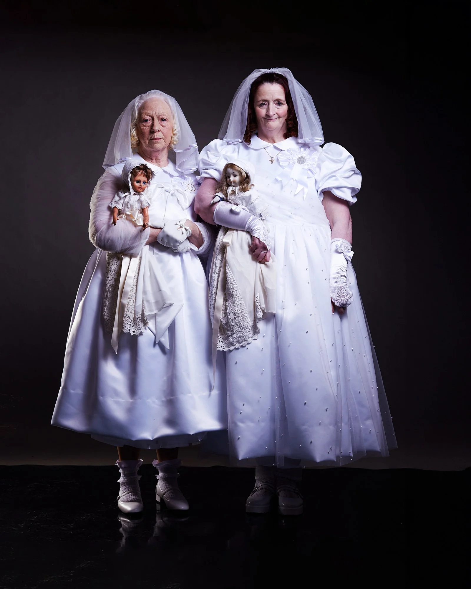 Advertising photo of two old women dressed as brides in white wedding gowns and veils, holding vintage dolls, stand against a dark background in a photo studio with professional lighting.
