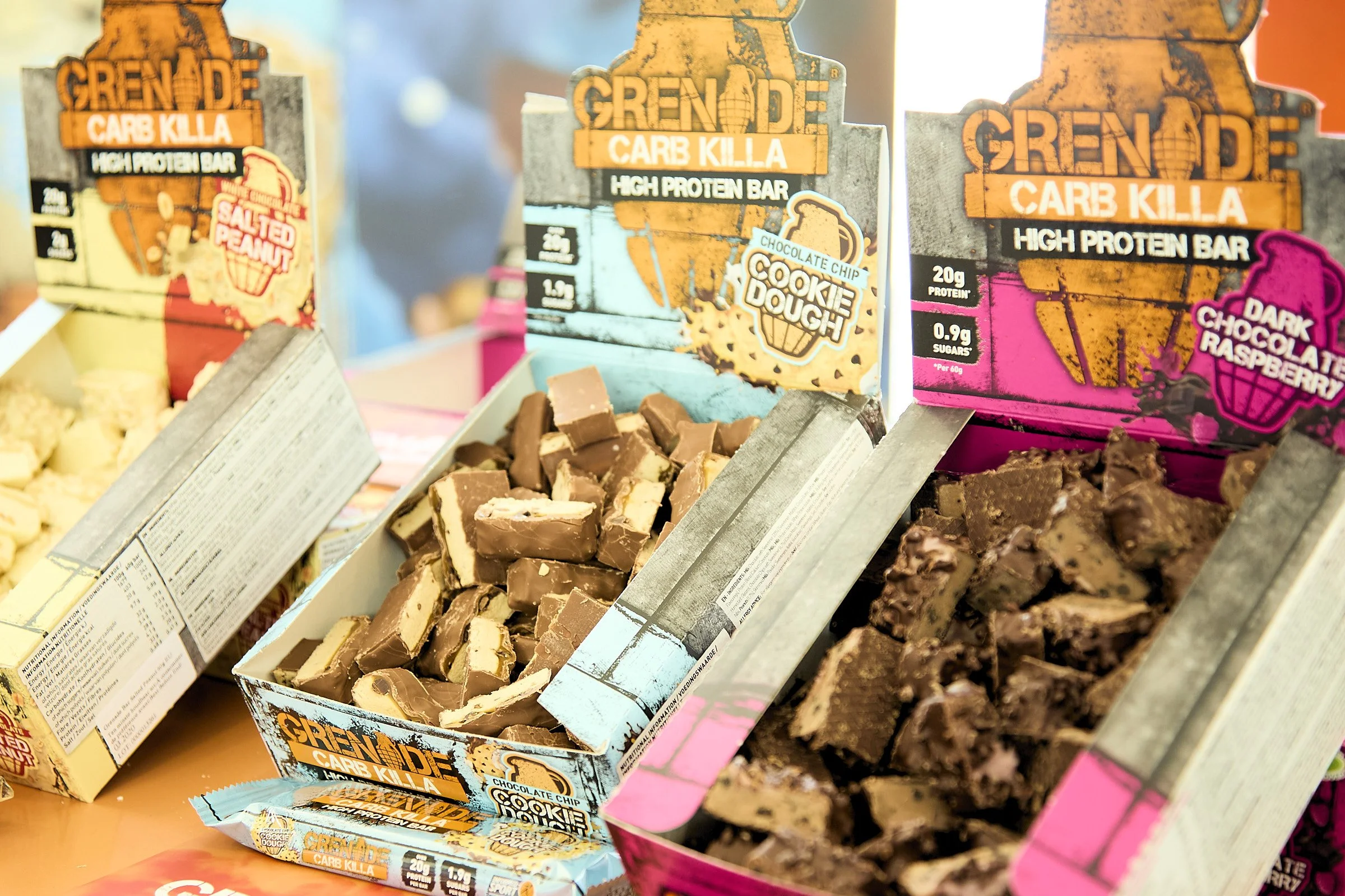 Three open boxes of high-protein snack bars with colorful packaging. The left box contains salted peanut flavor, the middle chocolate chip cookie dough flavor, and the right dark chocolate raspberry flavor.