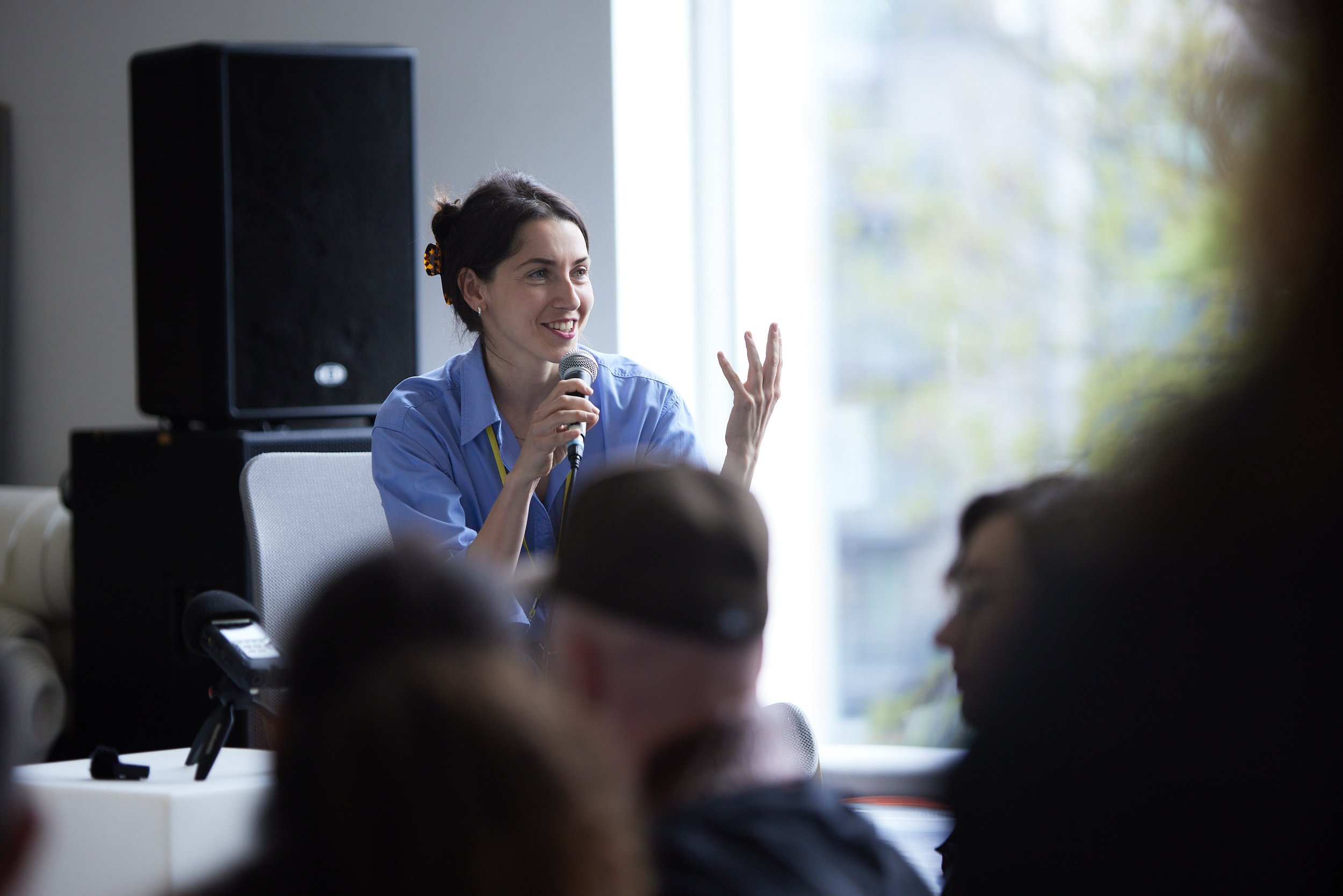 A woman in a blue shirt speaking into a microphone during a presentation or discussion, with an audience in front of her, in a room with large windows and natural light.