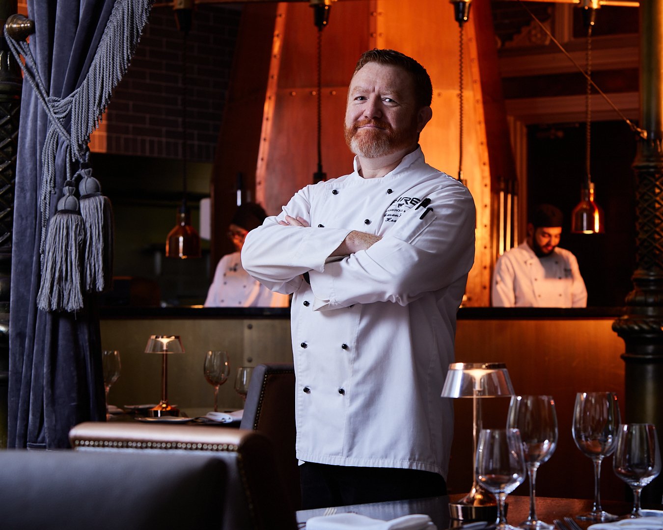 A male chef in a white chef's coat standing with arms crossed in a restaurant, with two other chefs working in the background, set in a warmly lit dining area.